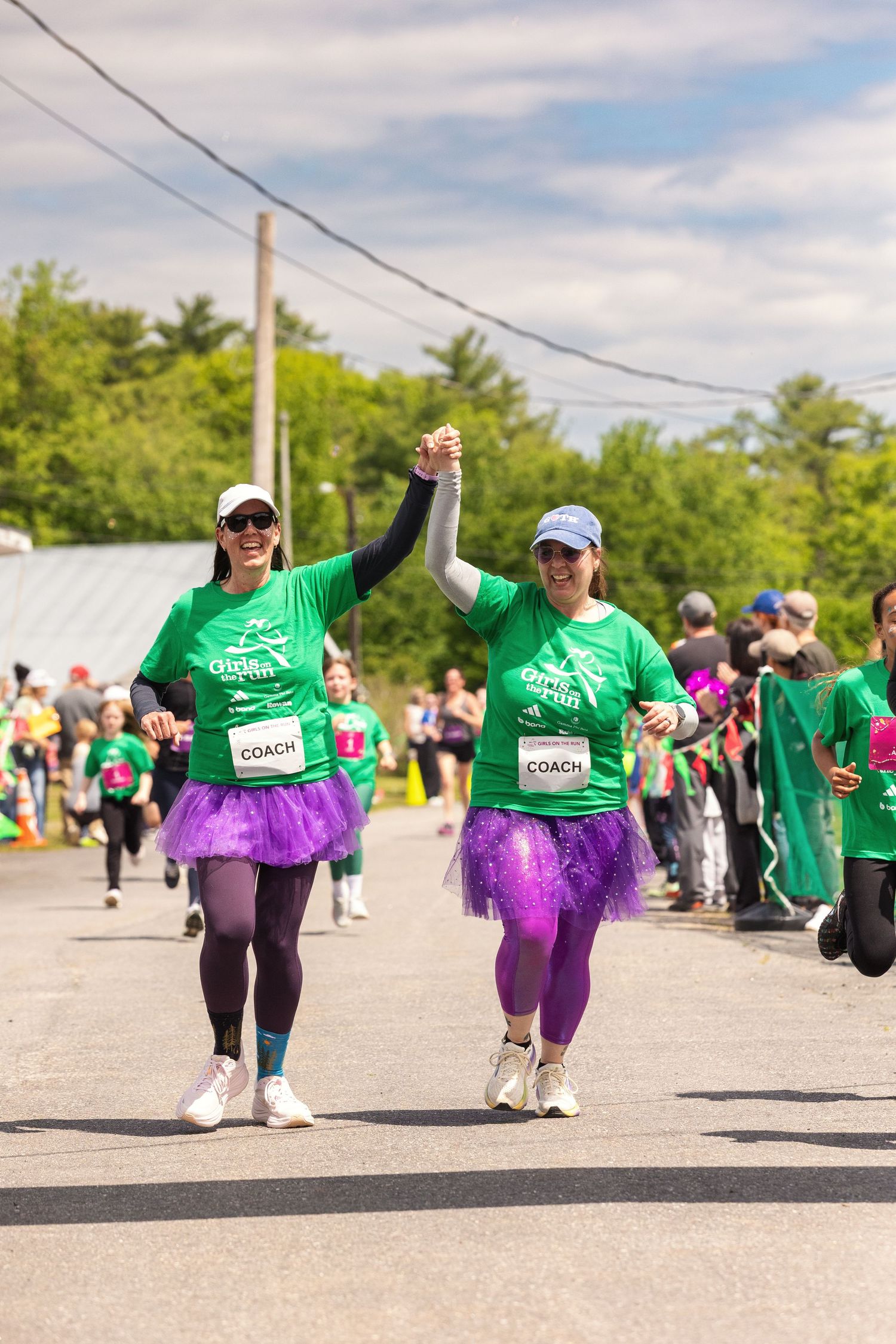 Two women coaches in green shirts and purple tutus hold hands while running a charity race outdoors.
