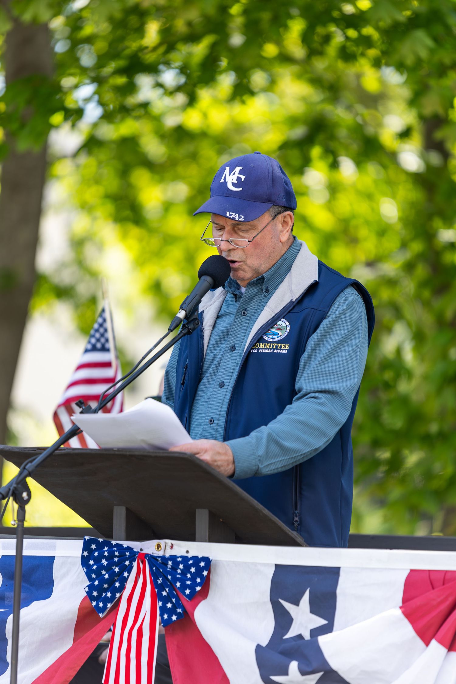 Elderly man in blue vest and cap reads speech at patriotic podium decorated with American flags outdoors.