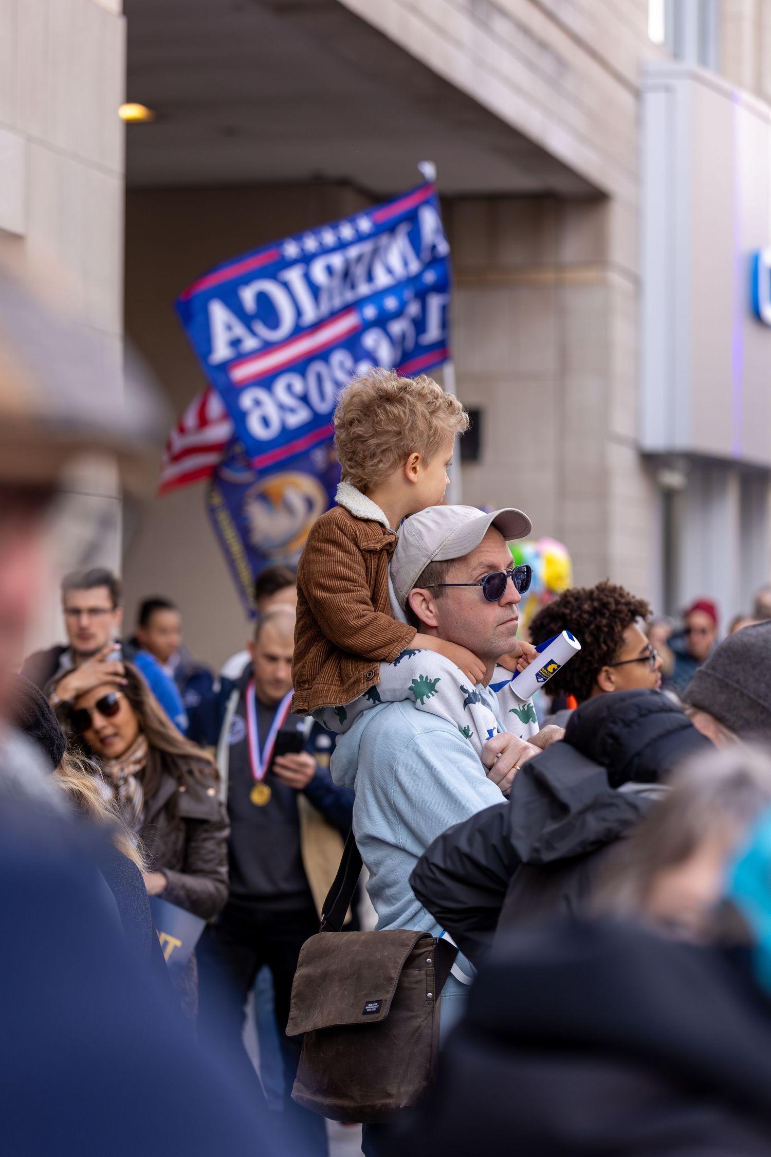 Man carries child on shoulders at a political rally with a Trump campaign flag visible in the crowd.
