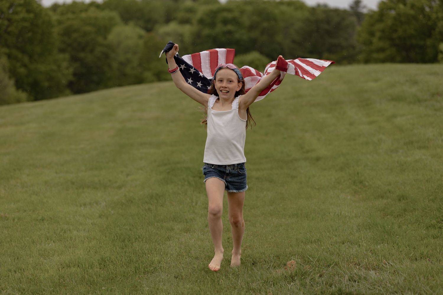 Young girl running barefoot on green grass, joyfully holding an American flag with both hands raised above her head.