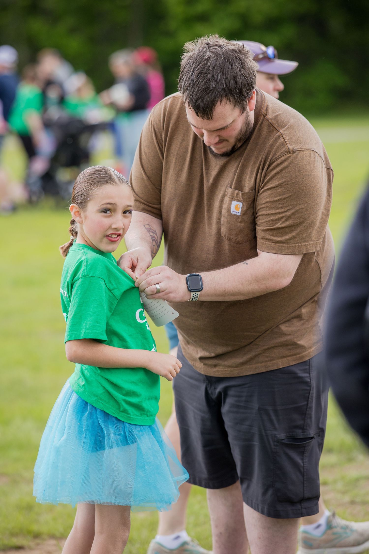 A man in a brown Carhartt shirt helps a young girl in a green tee and blue tutu at an outdoor event.