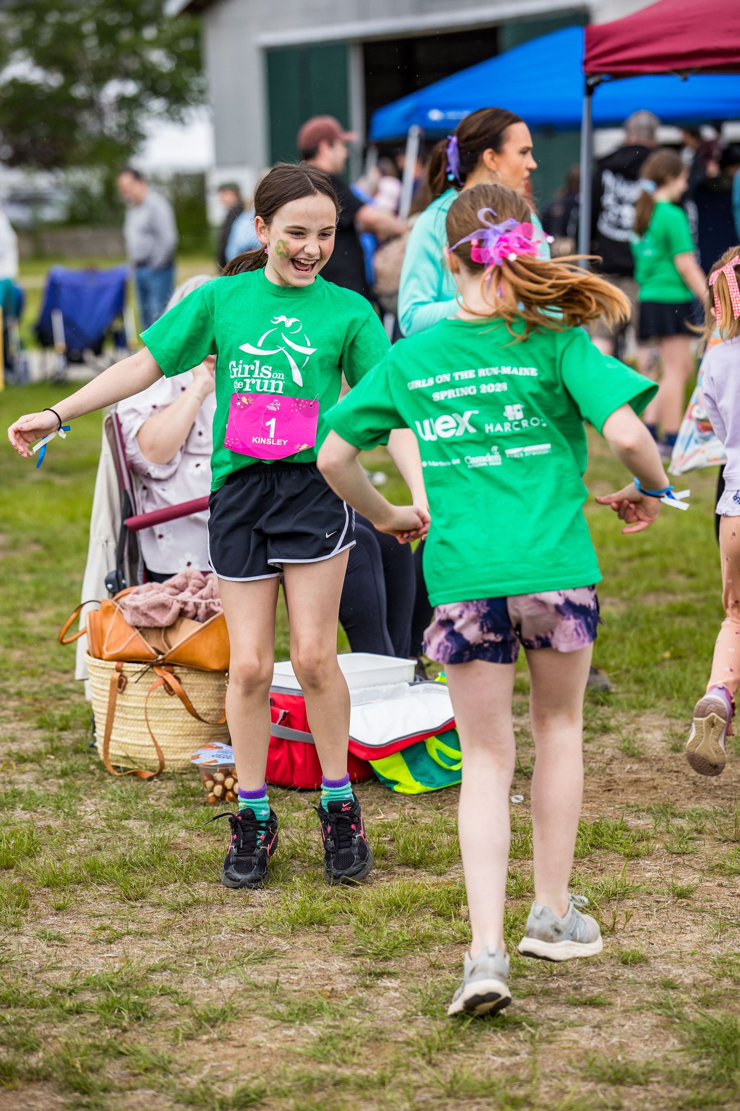 Two girls in green Girls on the Run shirts laugh and dance together at an outdoor 5K race event.