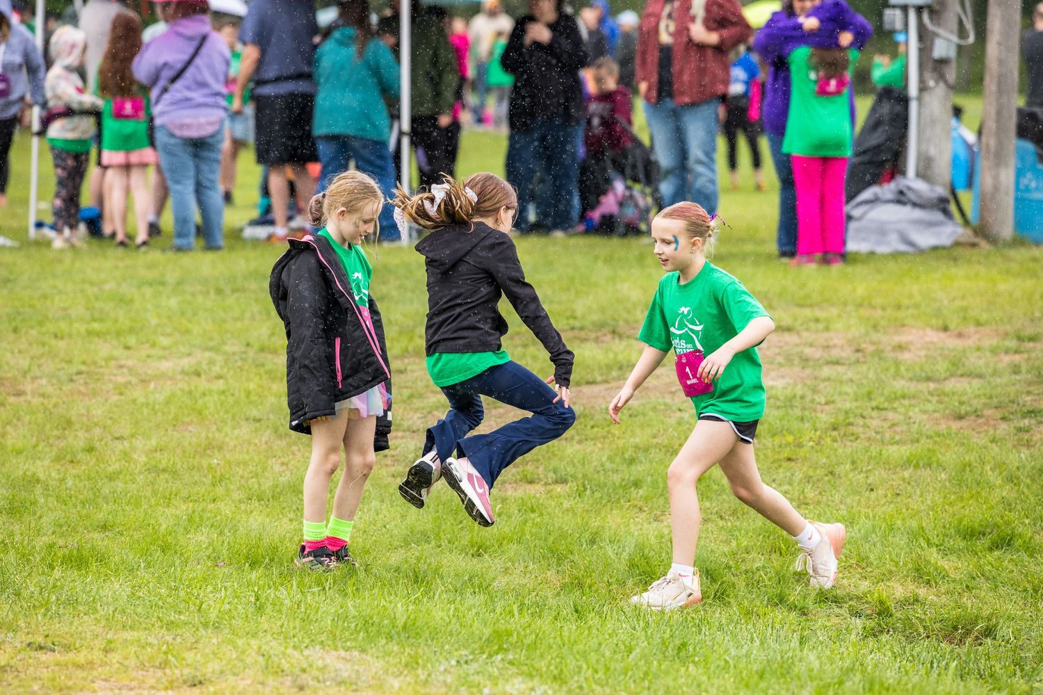 Children in green shirts play an outdoor game on grass at a school or community event on a cloudy day.