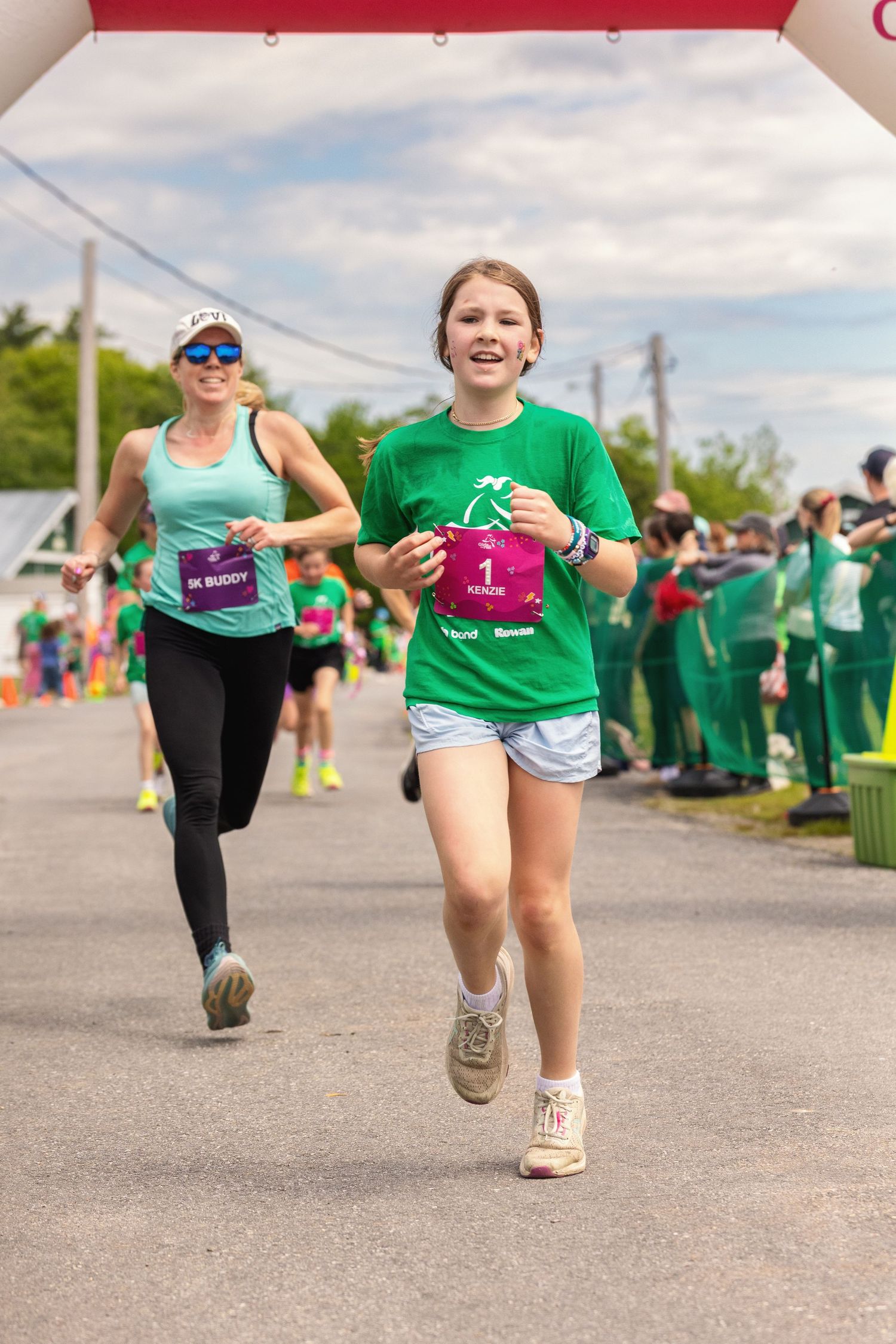 Young girl in green shirt running and smiling during a community race event with other participants in the background.