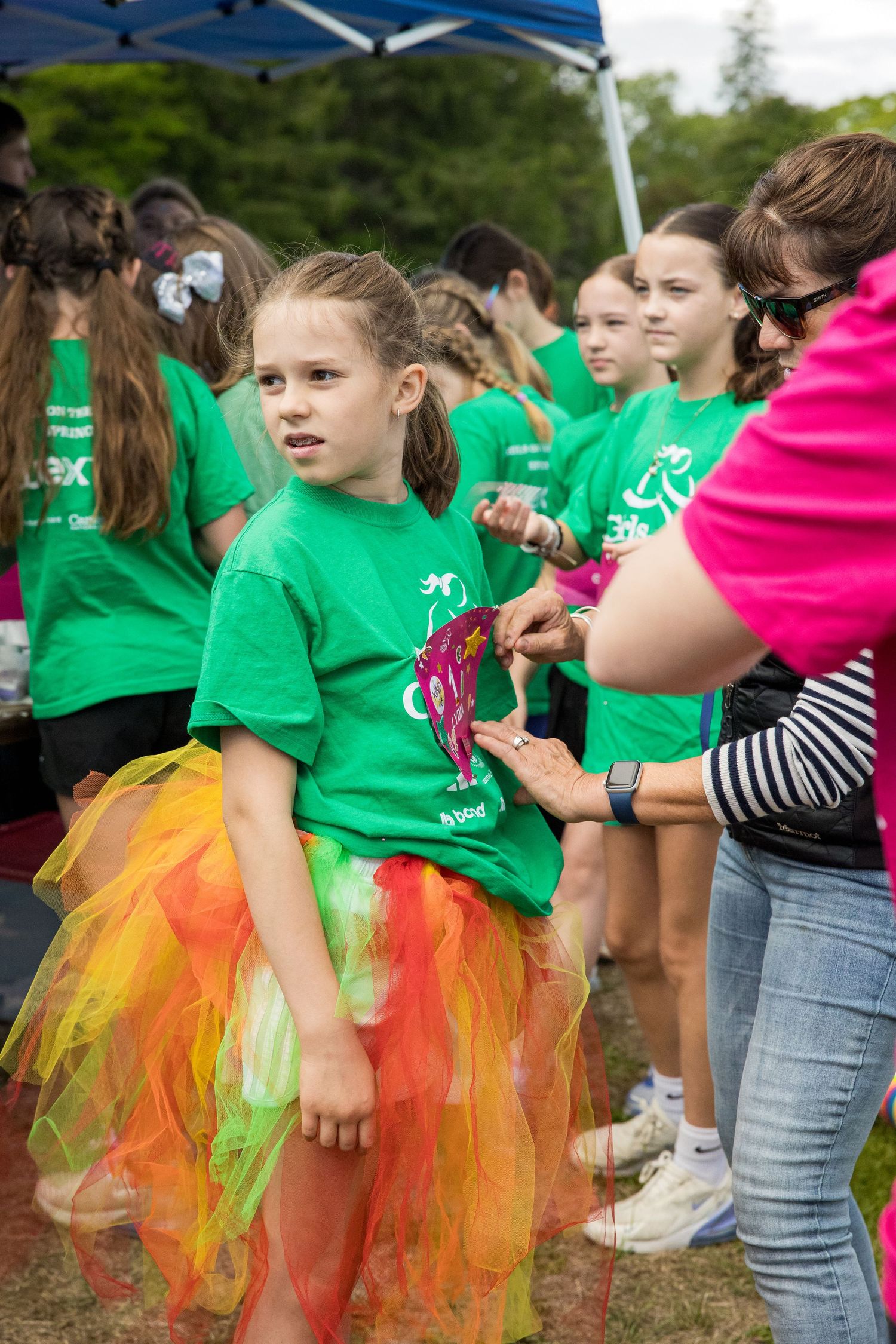 Girl in green shirt and colorful tutu skirt gets medal pinned at outdoor charity run event with group of kids.