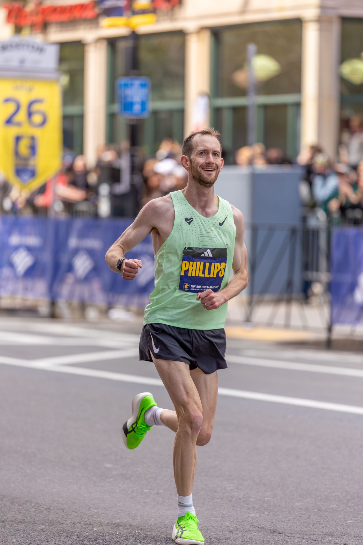 Runner Phillips competing in the Boston Marathon, wearing a green vest and black shorts, smiling near mile 26.
