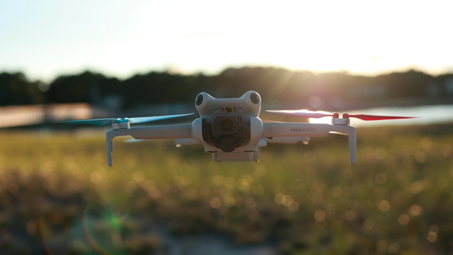 A DJI Mini drone hovers low over a sunlit grassy field near water during golden hour sunset.