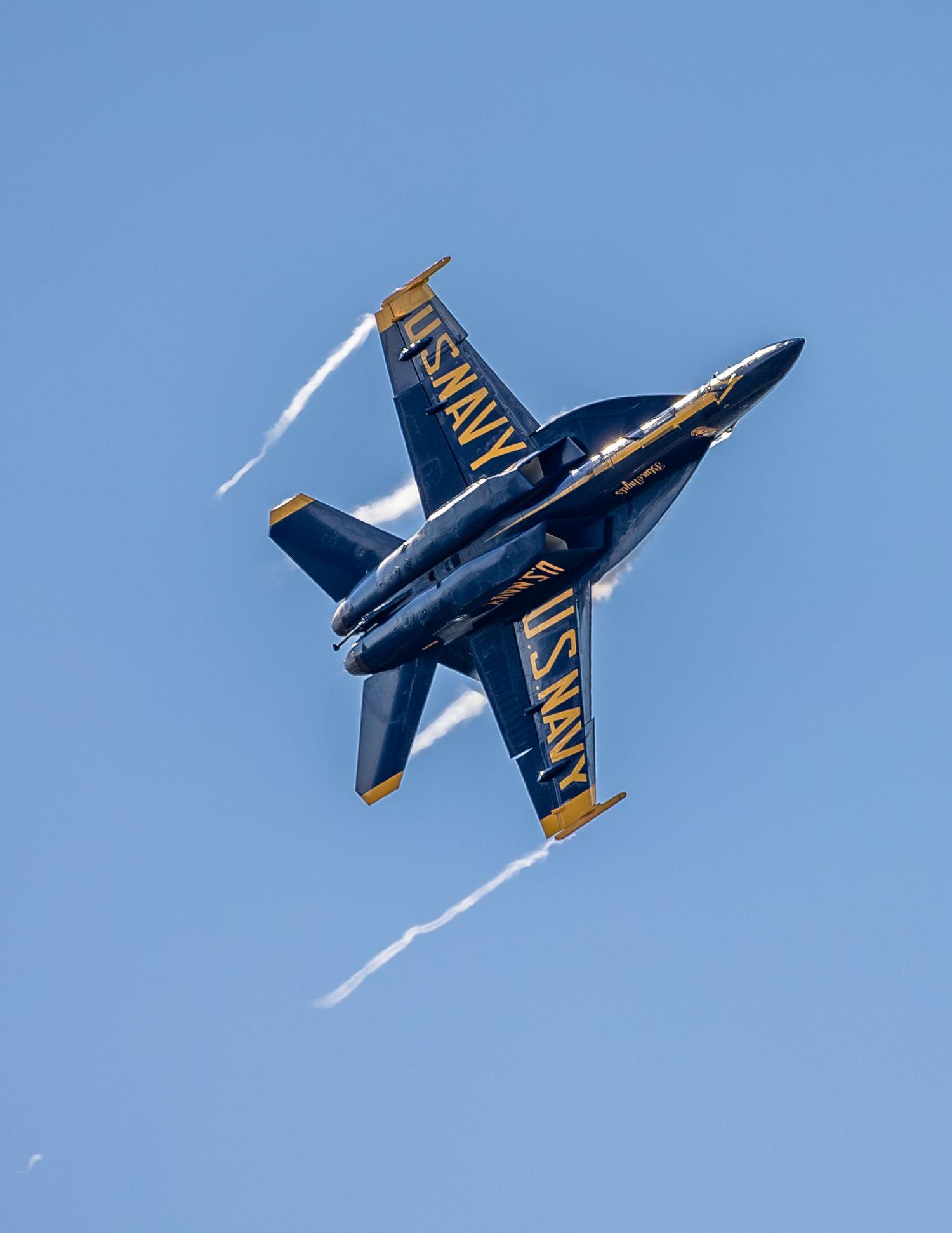 US Navy Blue Angels F/A-18 jet performing a dramatic inverted maneuver at an airshow against a clear blue sky.