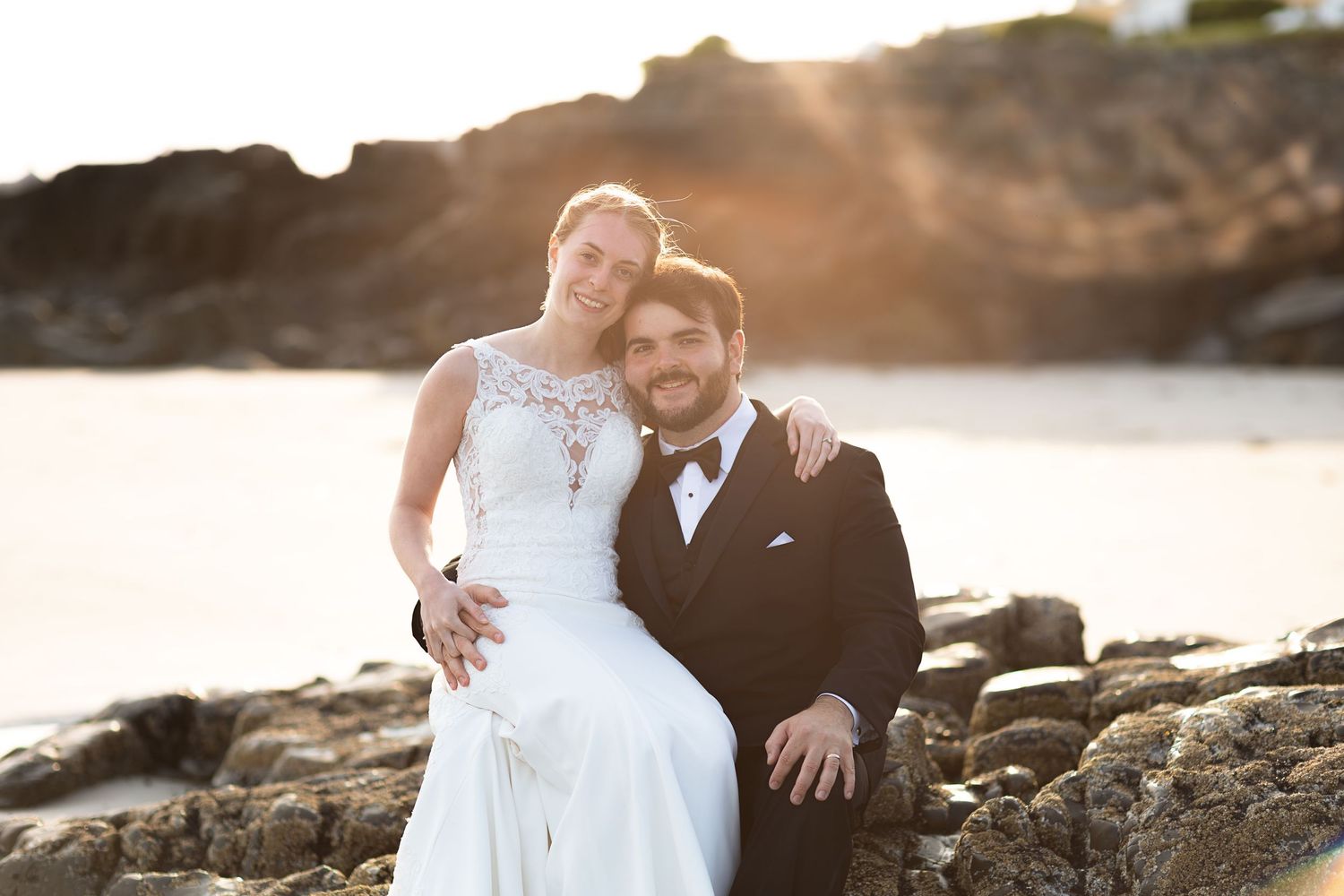 Bride and groom sitting on rocky beach at sunset, smiling together in wedding attire.