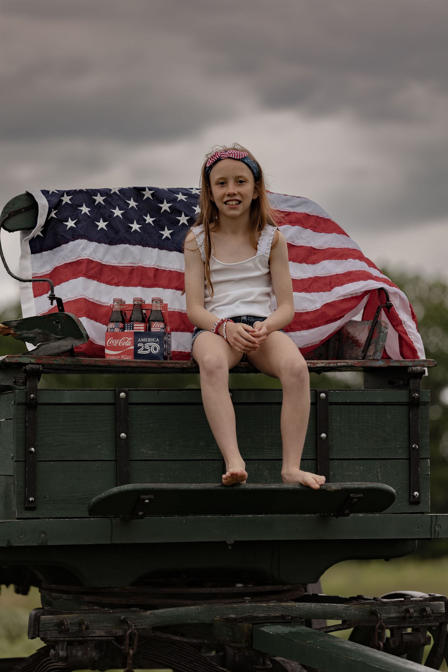Young girl sitting on green military vehicle with American flag backdrop and Coca-Cola bottles on cloudy day.