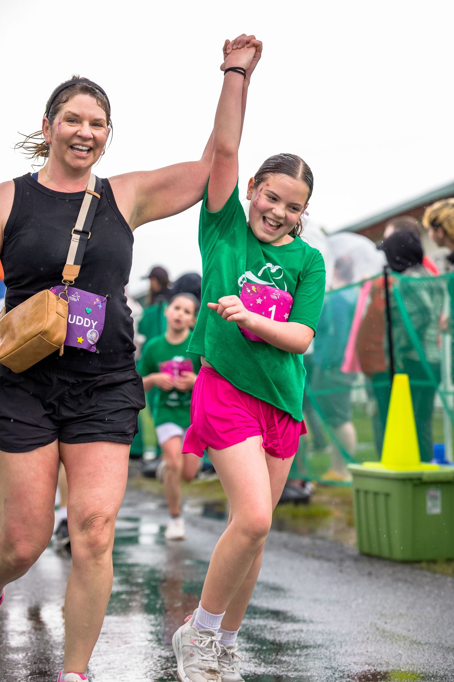Girl in green shirt celebrates finishing a fun run race with adult buddy, both smiling on wet pavement.