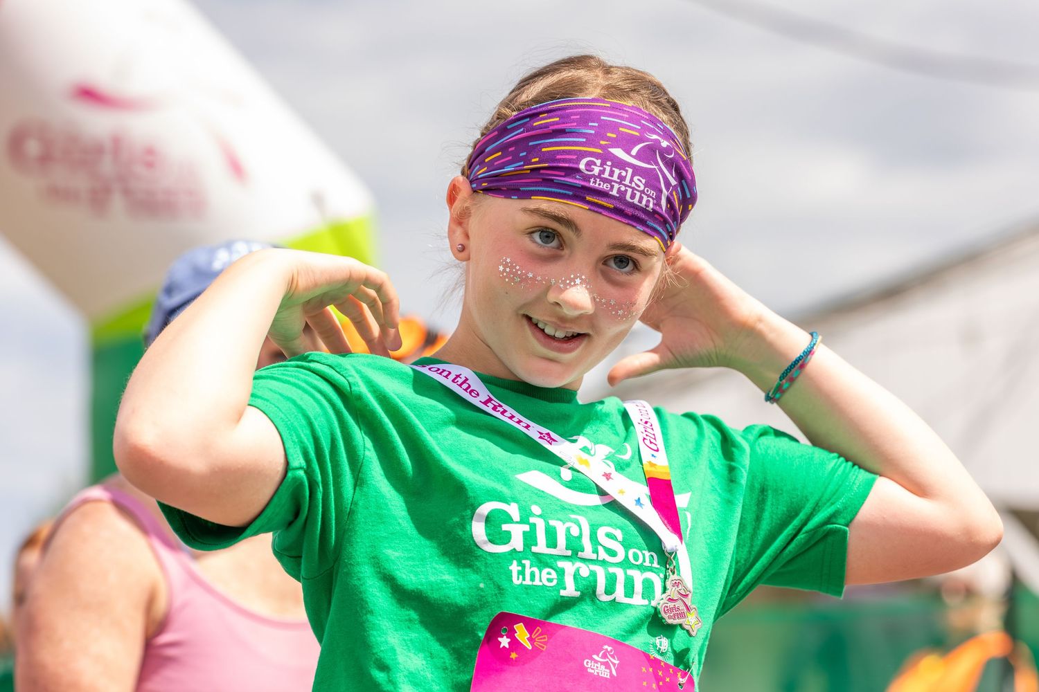 Young girl wearing a green Girls on the Run t-shirt and purple headband smiling at a running event.