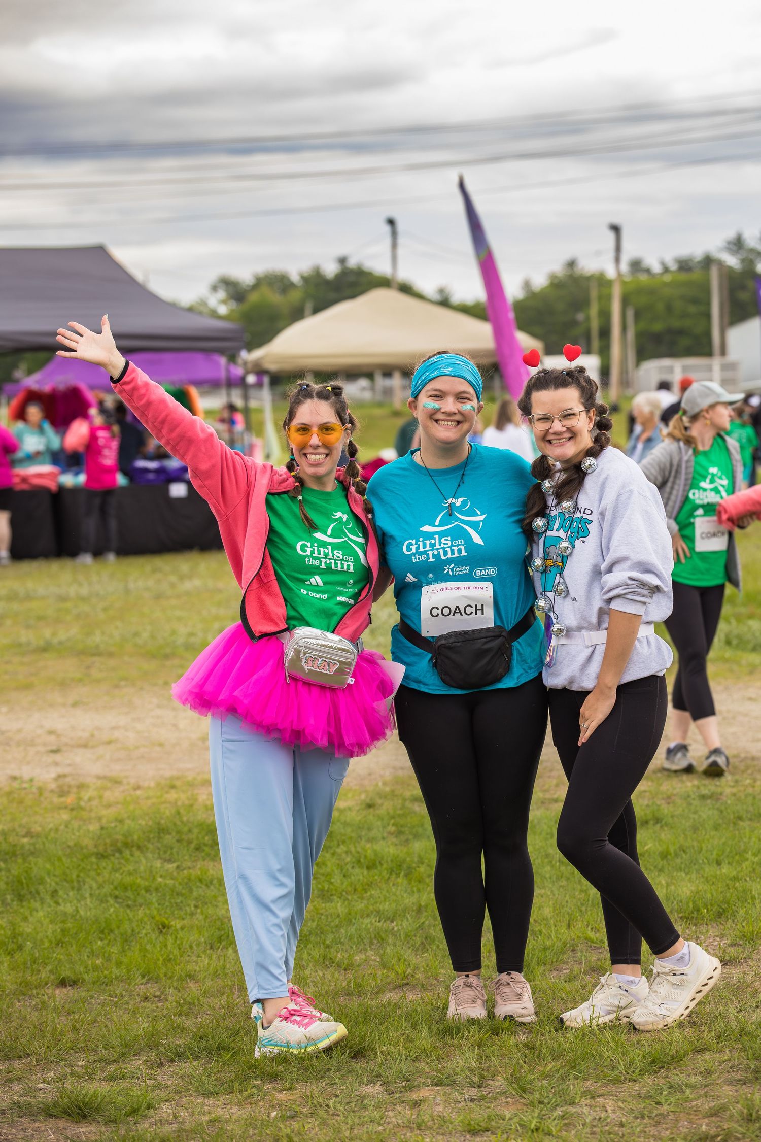 Three women in colorful outfits pose together at a Girls on the Run 5K event on a grassy field.