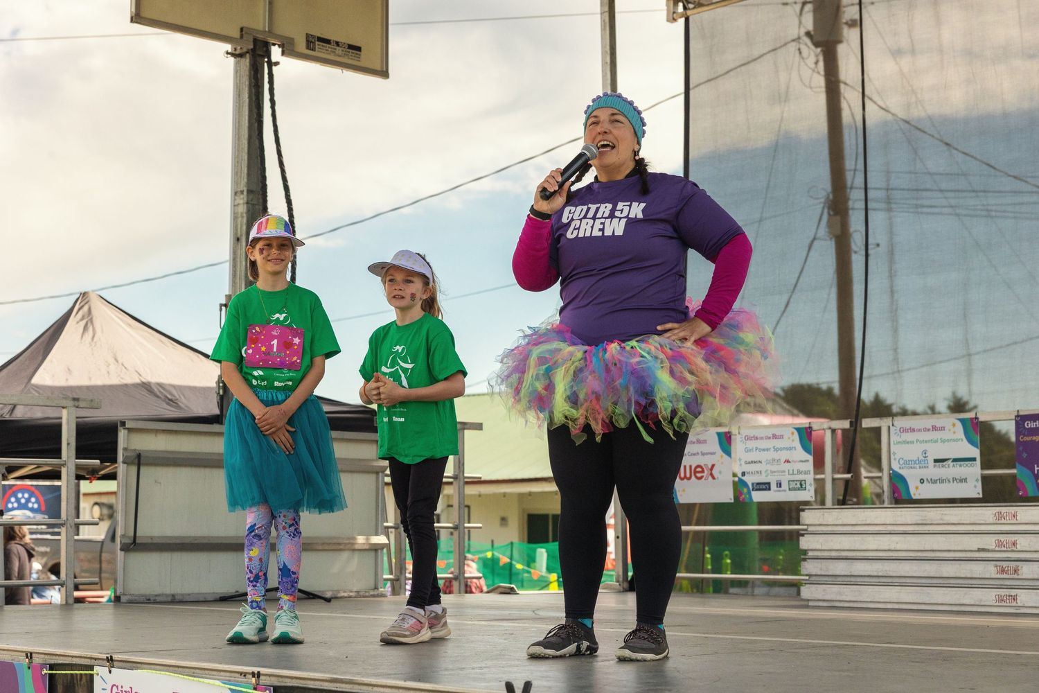 Three women on stage at a charity run event, wearing colorful tutus and green shirts, with one speaking into a microphone.