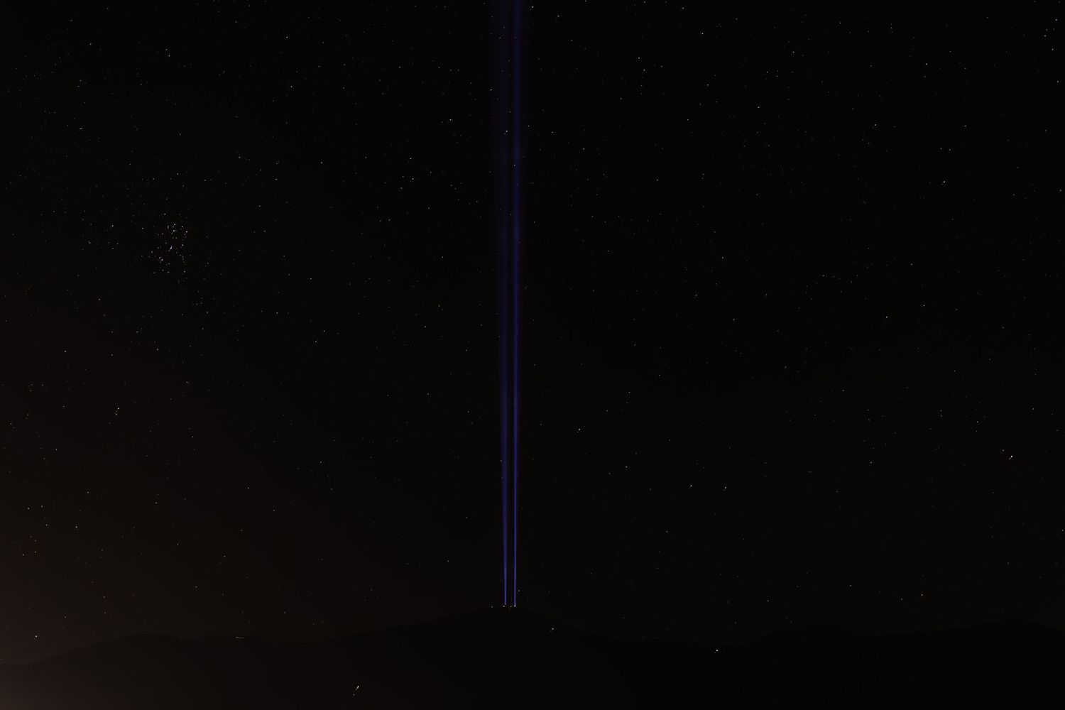 Blue laser beams streak upward into a dark night sky filled with faint stars in a long exposure photograph.