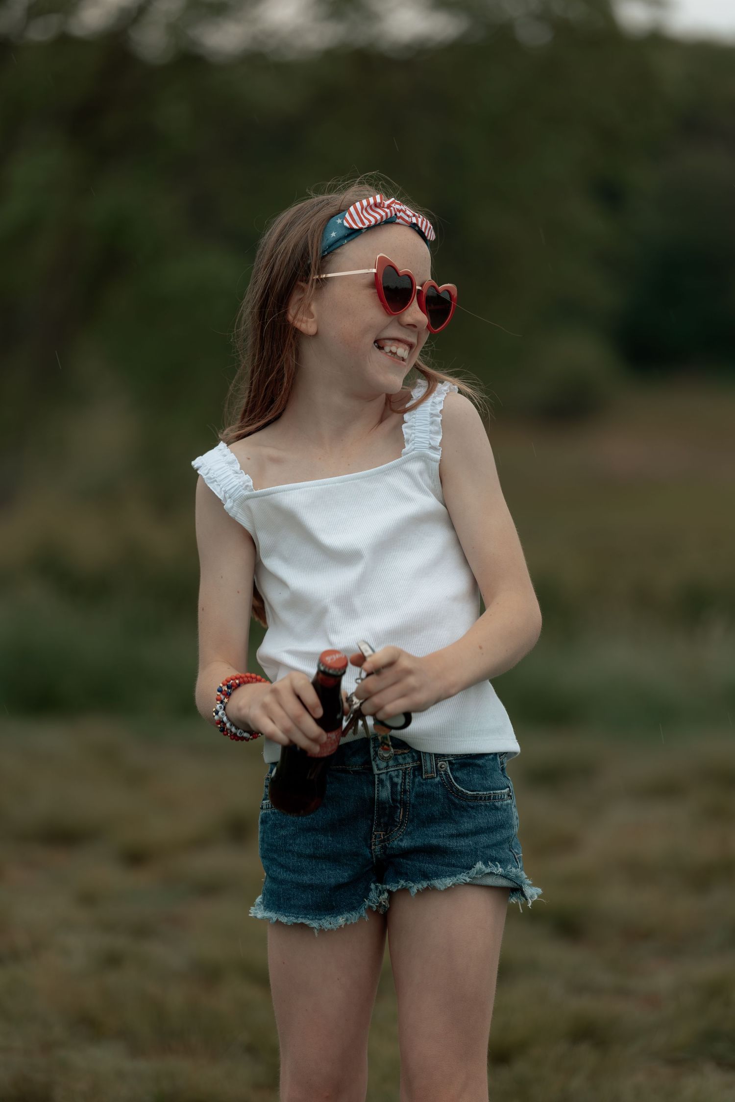 Smiling girl in white tank top, denim shorts, red heart sunglasses, and patriotic headband holding a bottle outdoors.