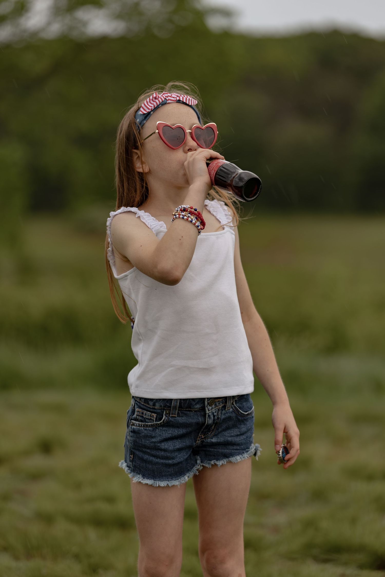 Young girl in heart sunglasses and denim shorts drinking from a bottle outdoors in a green field.