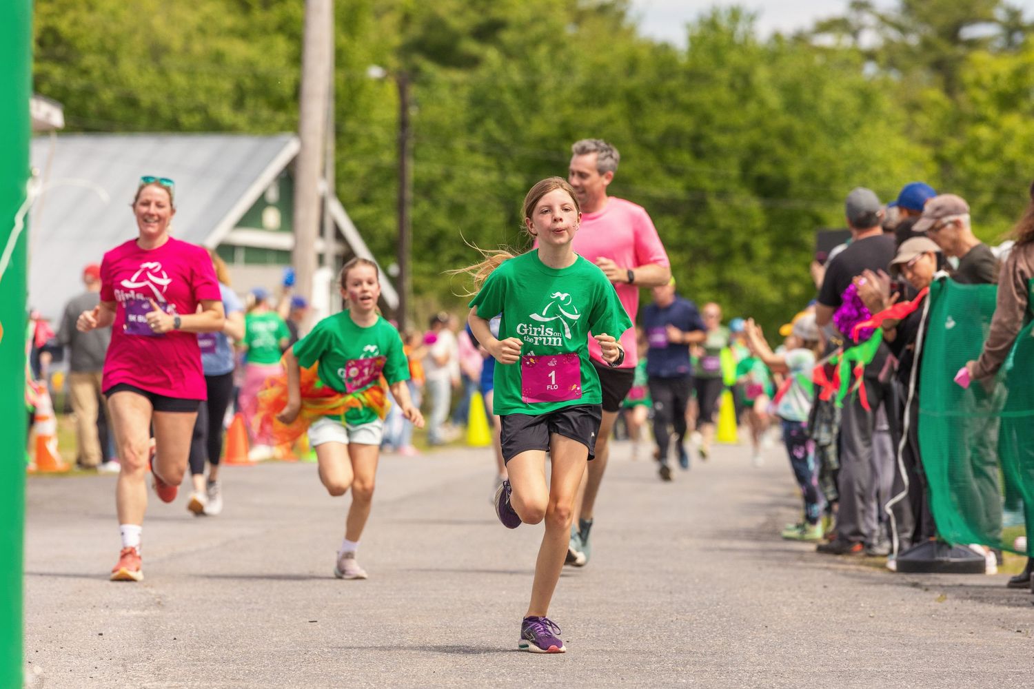 Children and adults racing in a fun run event on a sunny day, wearing colorful shirts with cheering crowd.