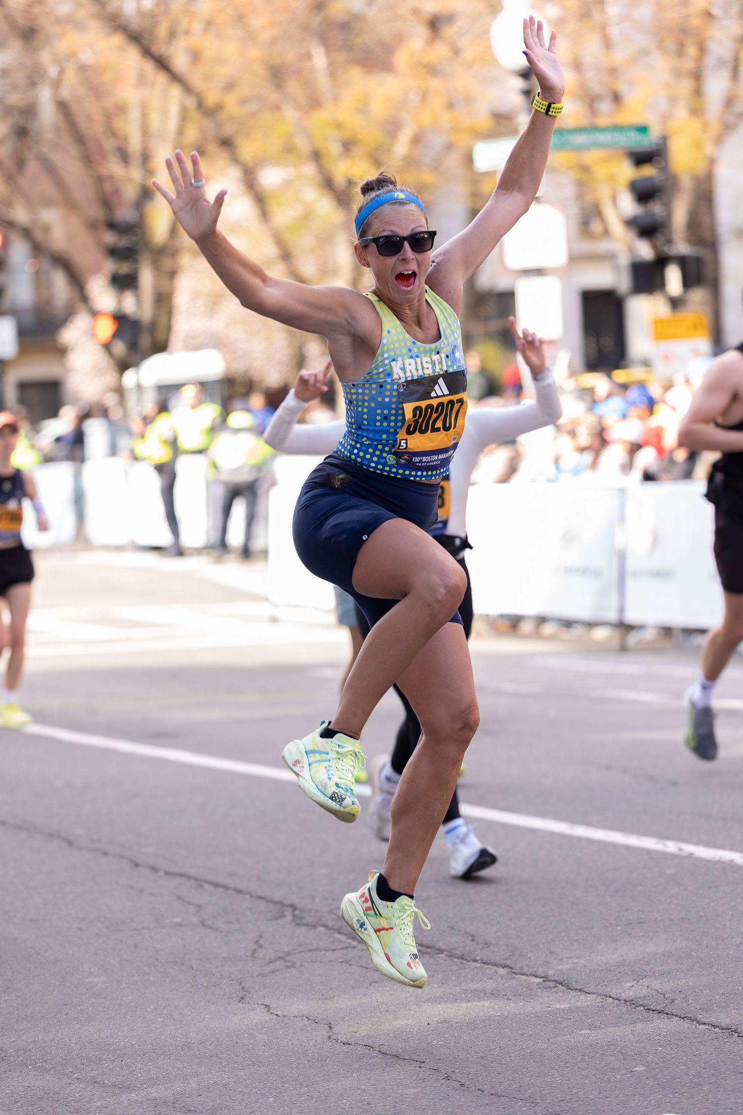Excited female marathon runner Kristin leaps joyfully mid-race wearing bib 30207 at Boston Marathon.