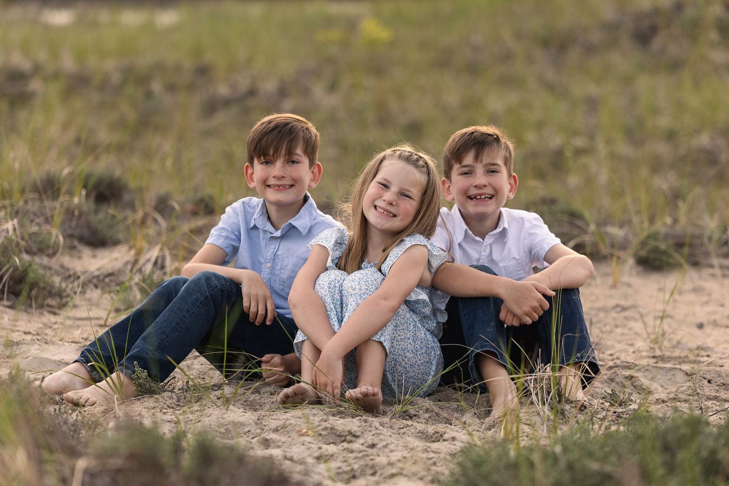 Three smiling children sit together on sandy ground in a field, posing for an outdoor family portrait photo.