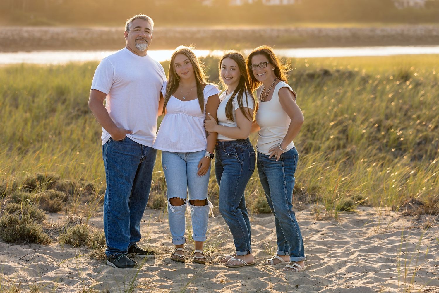 Family wearing jeans and white shirts walks together on beach path during golden hour sunset.
