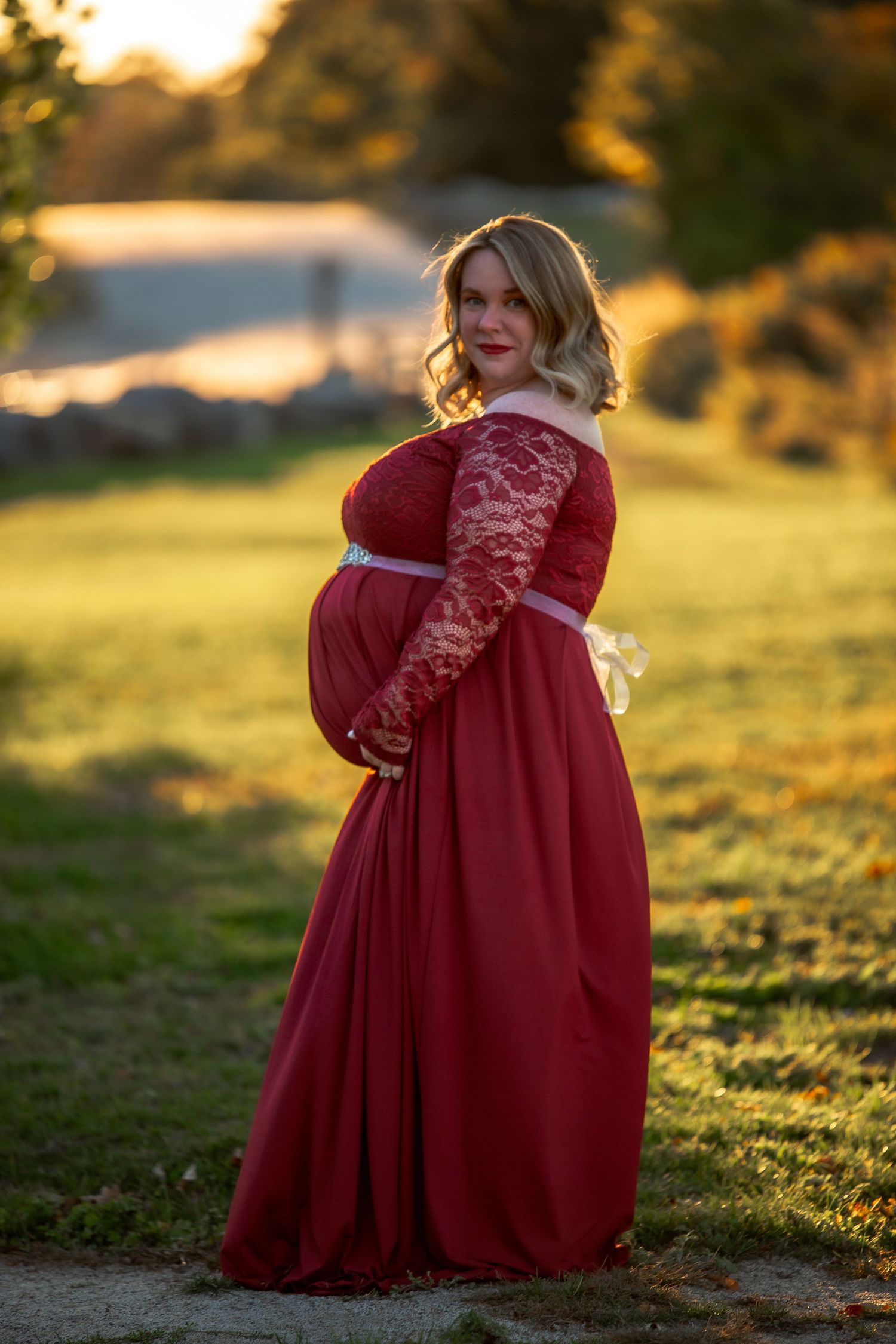 A figure in a flowing red lace dress poses gracefully in a sunlit field during golden hour.