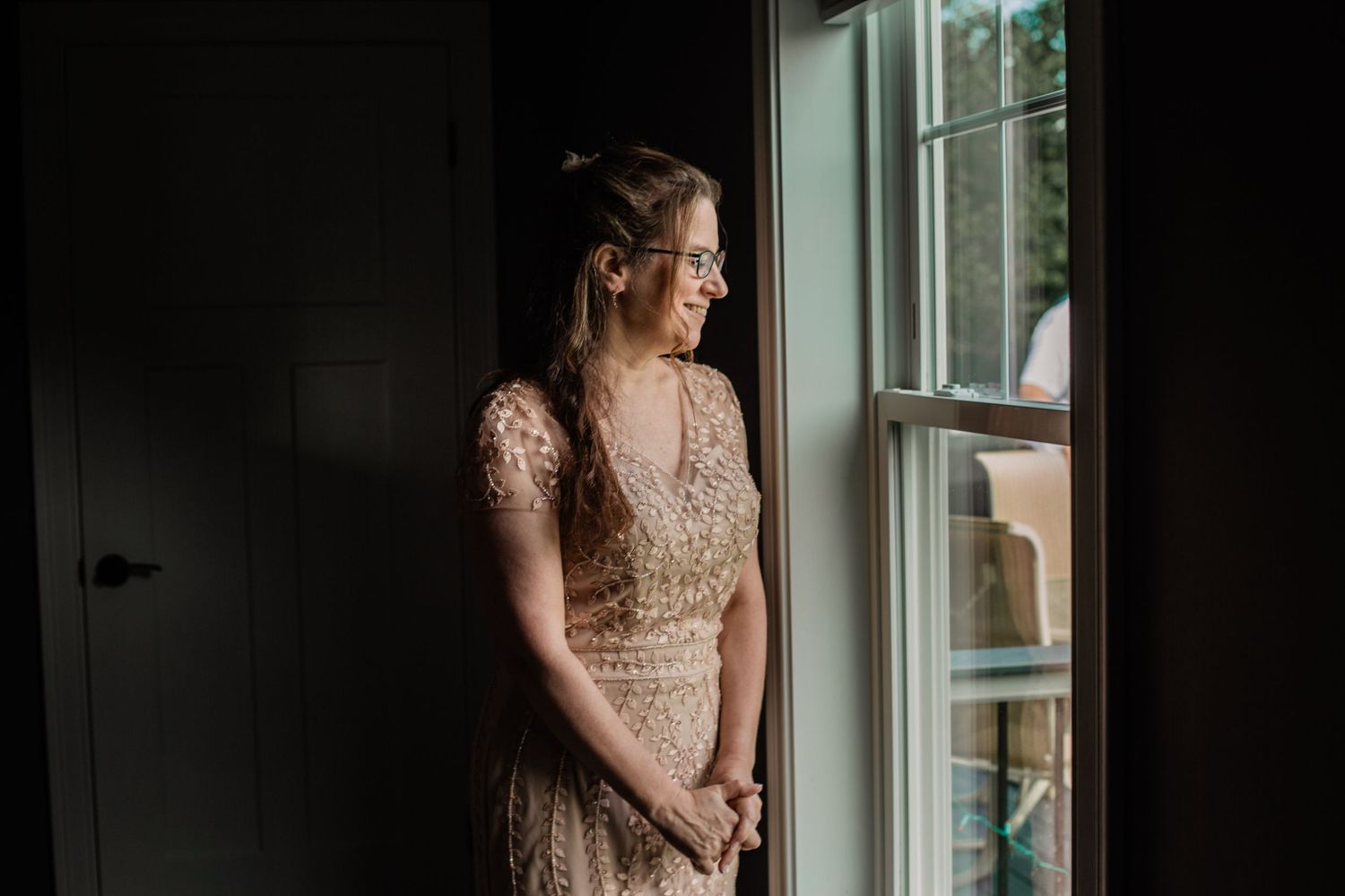 Bride in a vintage-inspired lace wedding gown poses elegantly by sunlit window in intimate bridal portrait.