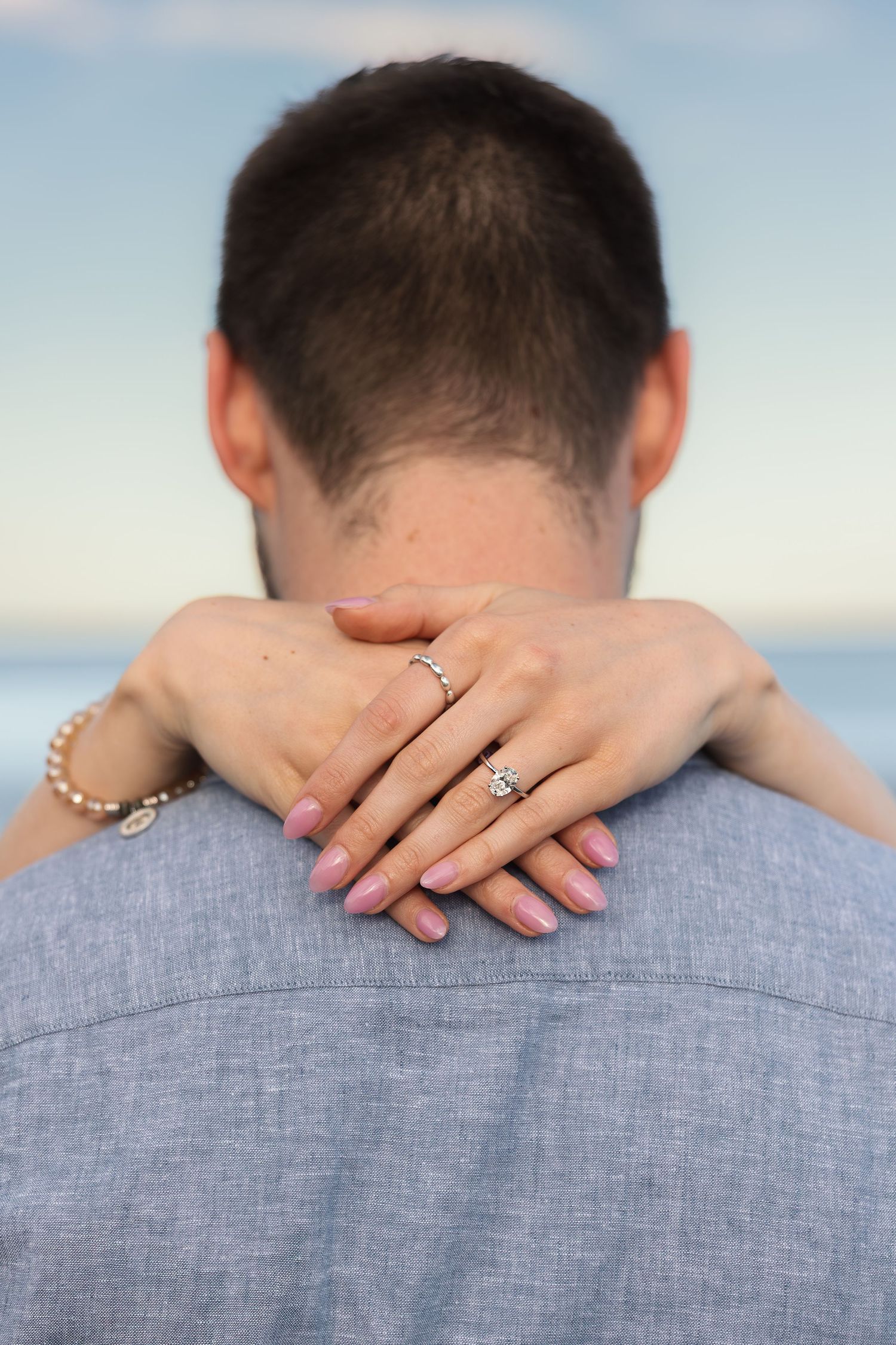 Woman with engagement ring and pink nails embracing a man from behind, with ocean in the background.
