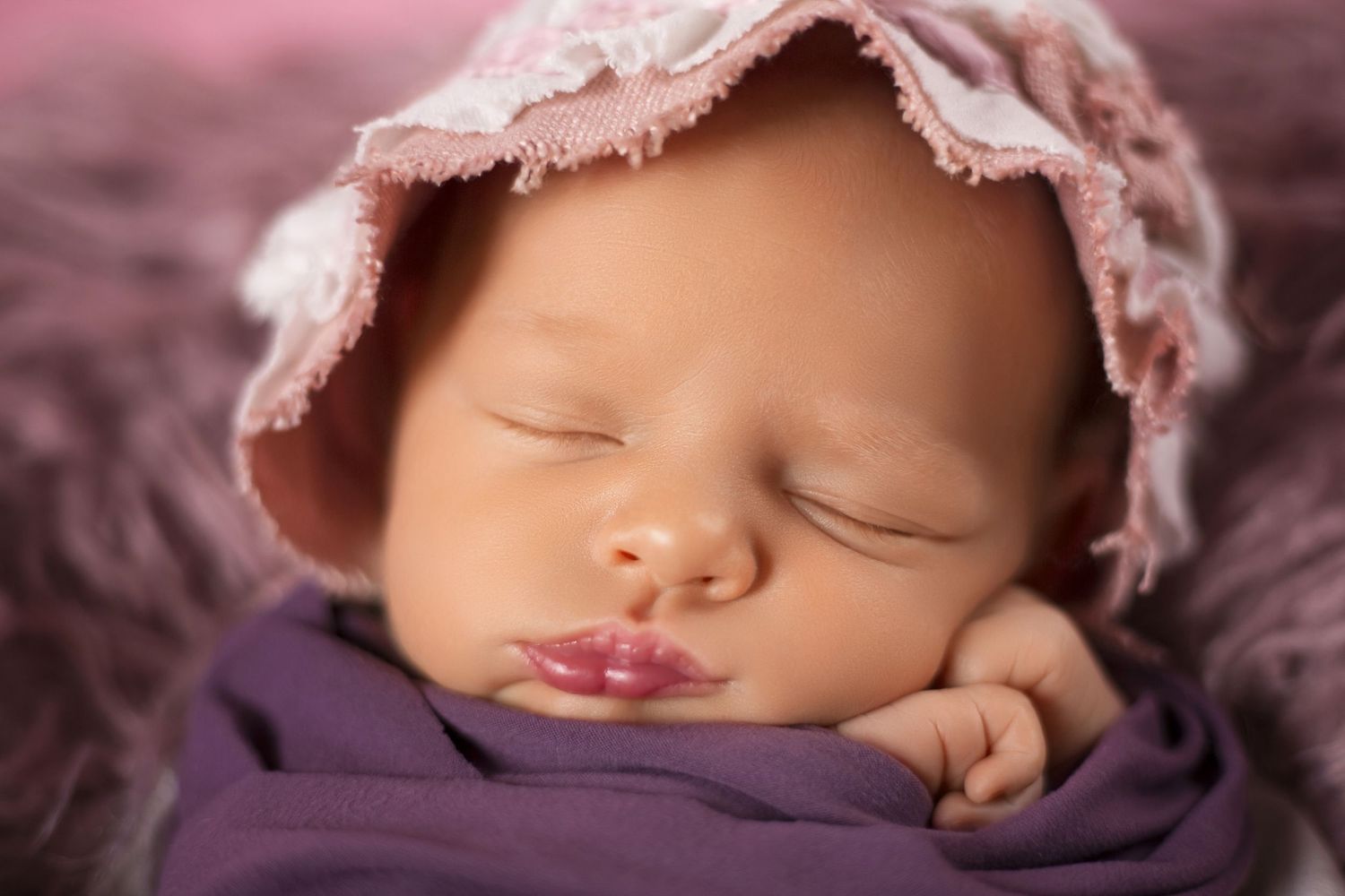 Peaceful newborn baby sleeping in purple wrap wearing a delicate pink lace bonnet on a soft lavender background.