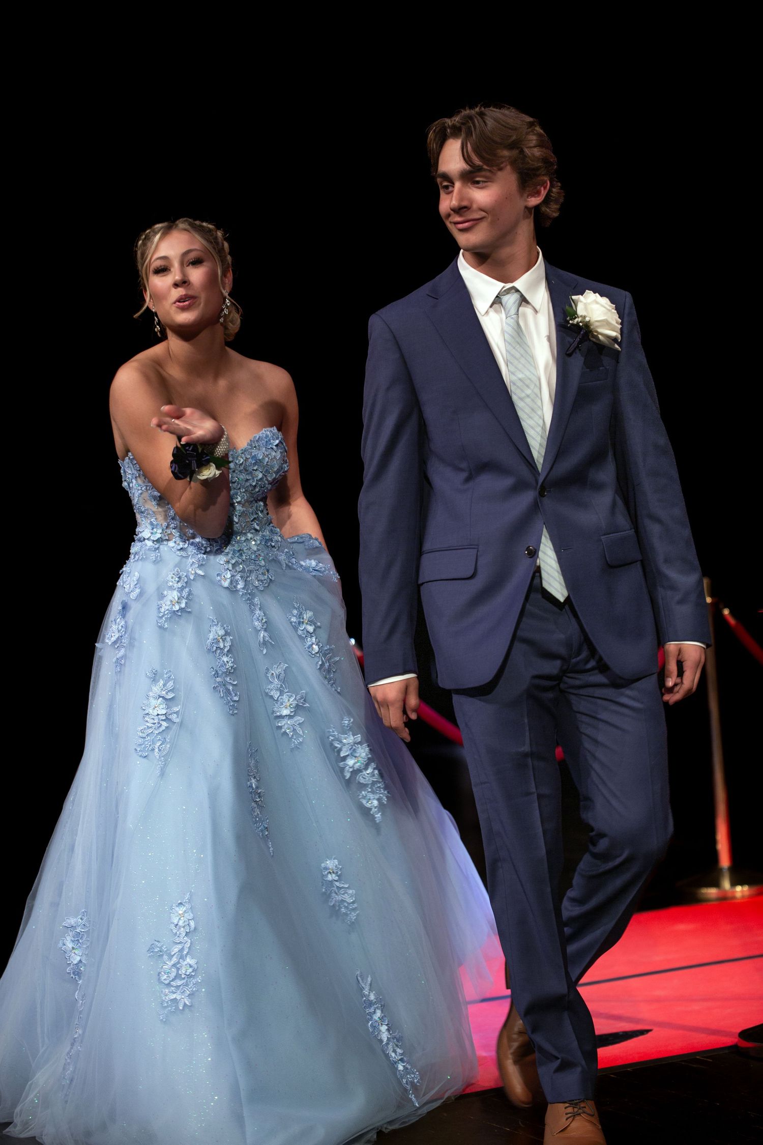 A young couple in formal attire, woman in blue gown and man in navy suit, walking a red carpet at prom.