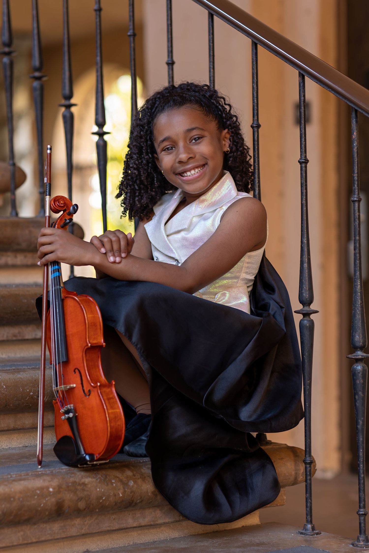 Smiling young girl in formal attire sitting on stairs holding a violin and bow.