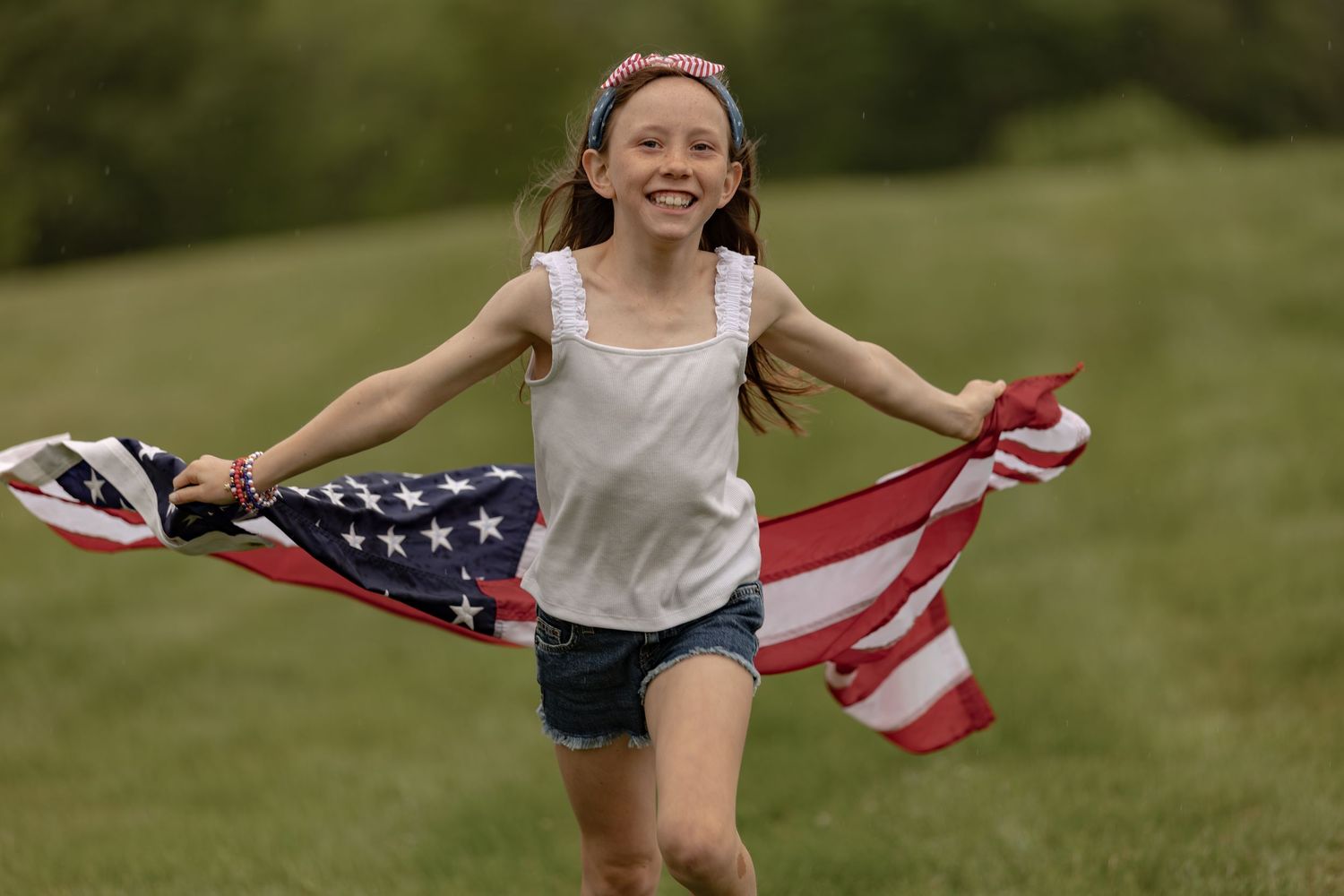 Smiling girl running through green field holding American flag, wearing white tank top and denim shorts.