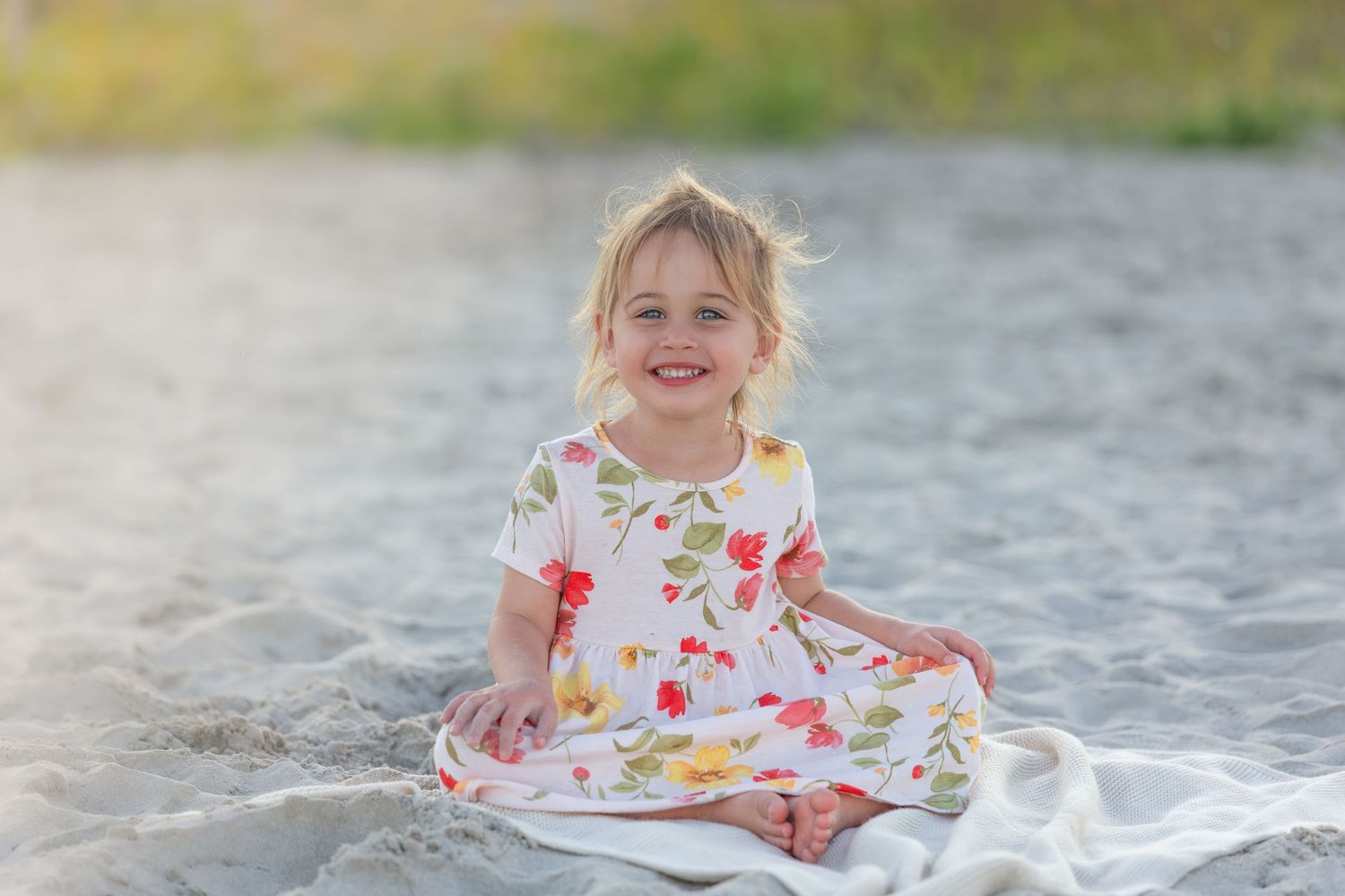Smiling toddler girl in floral dress sitting on sandy beach blanket outdoors.