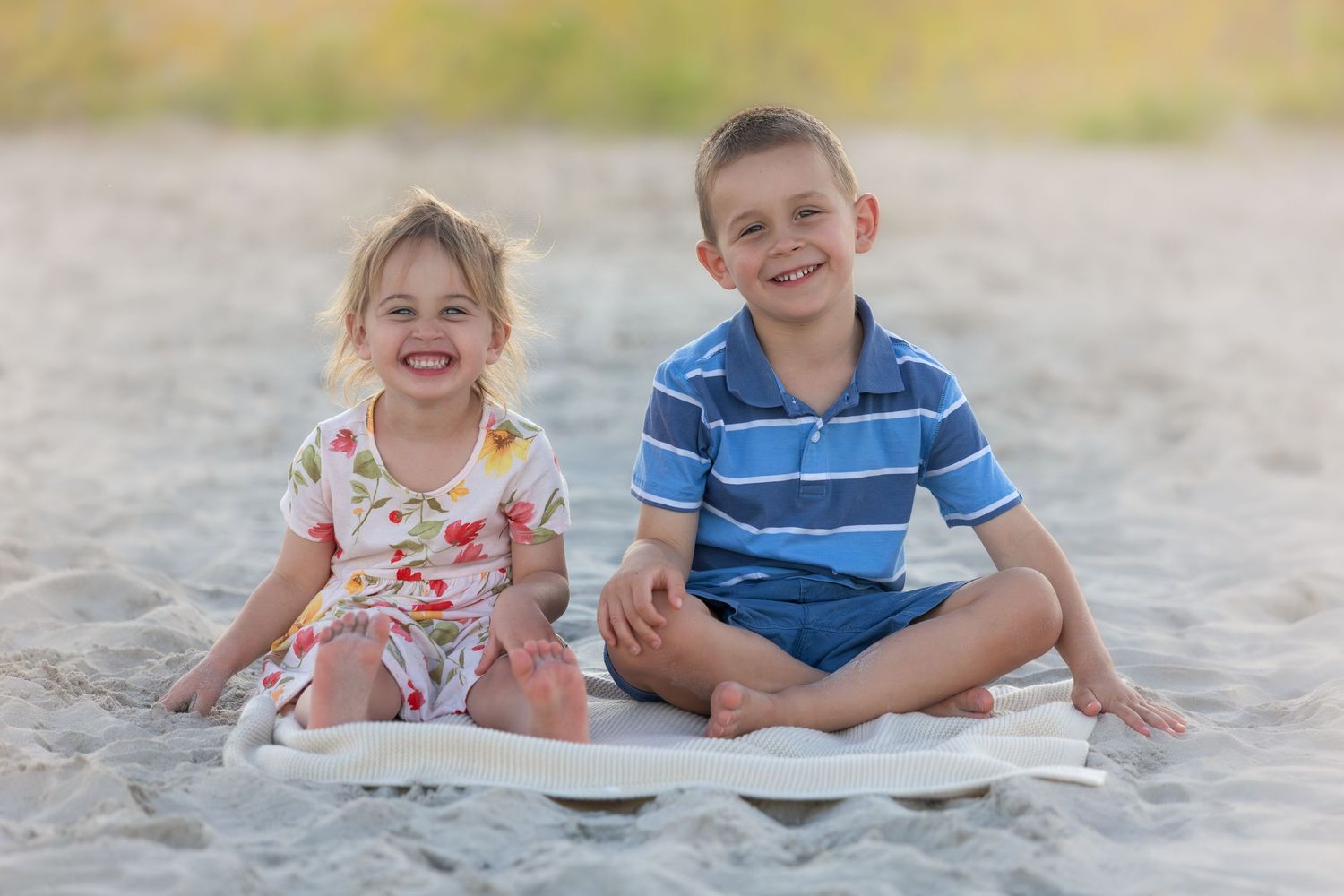 Two happy children sitting on a white blanket on a sandy beach, smiling outdoors on a sunny day.