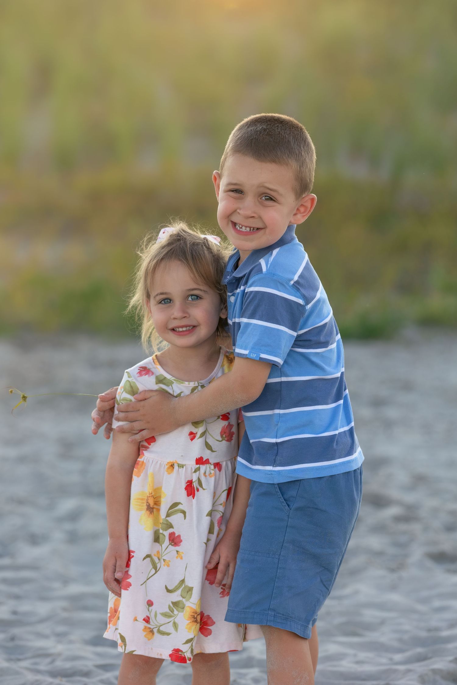 Two smiling children, a boy and girl, hugging on a sandy beach at golden hour.