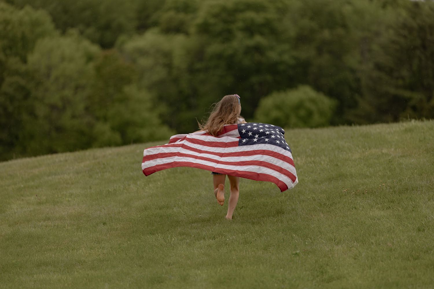 A young girl runs across a green hillside while holding an American flag that billows behind her.