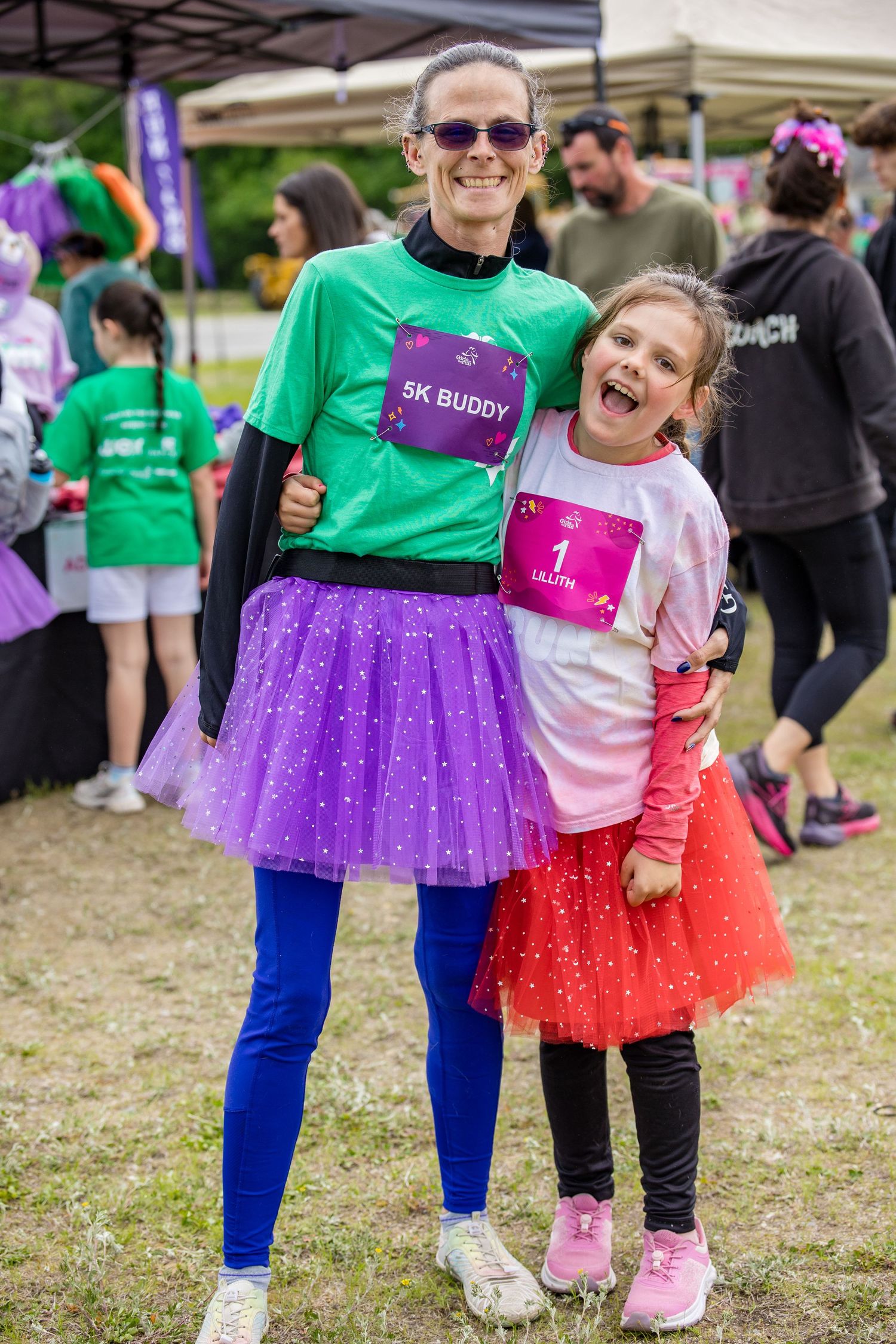 Woman and girl in colorful tutus pose together at a 5K fun run charity race event outdoors.