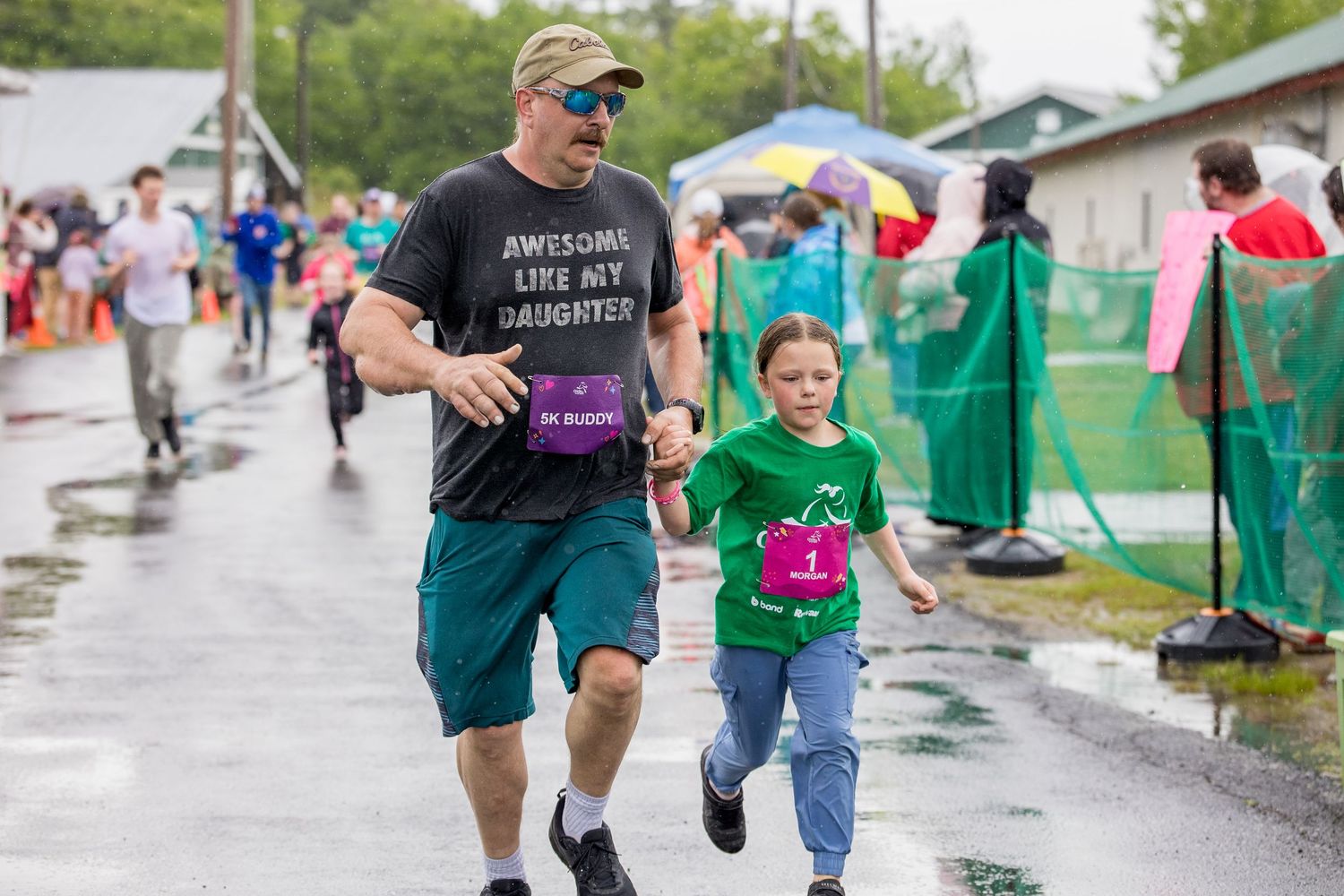 Man and girl running hand-in-hand in rainy charity race, man wearing 'Awesome Like My Daughter' shirt.