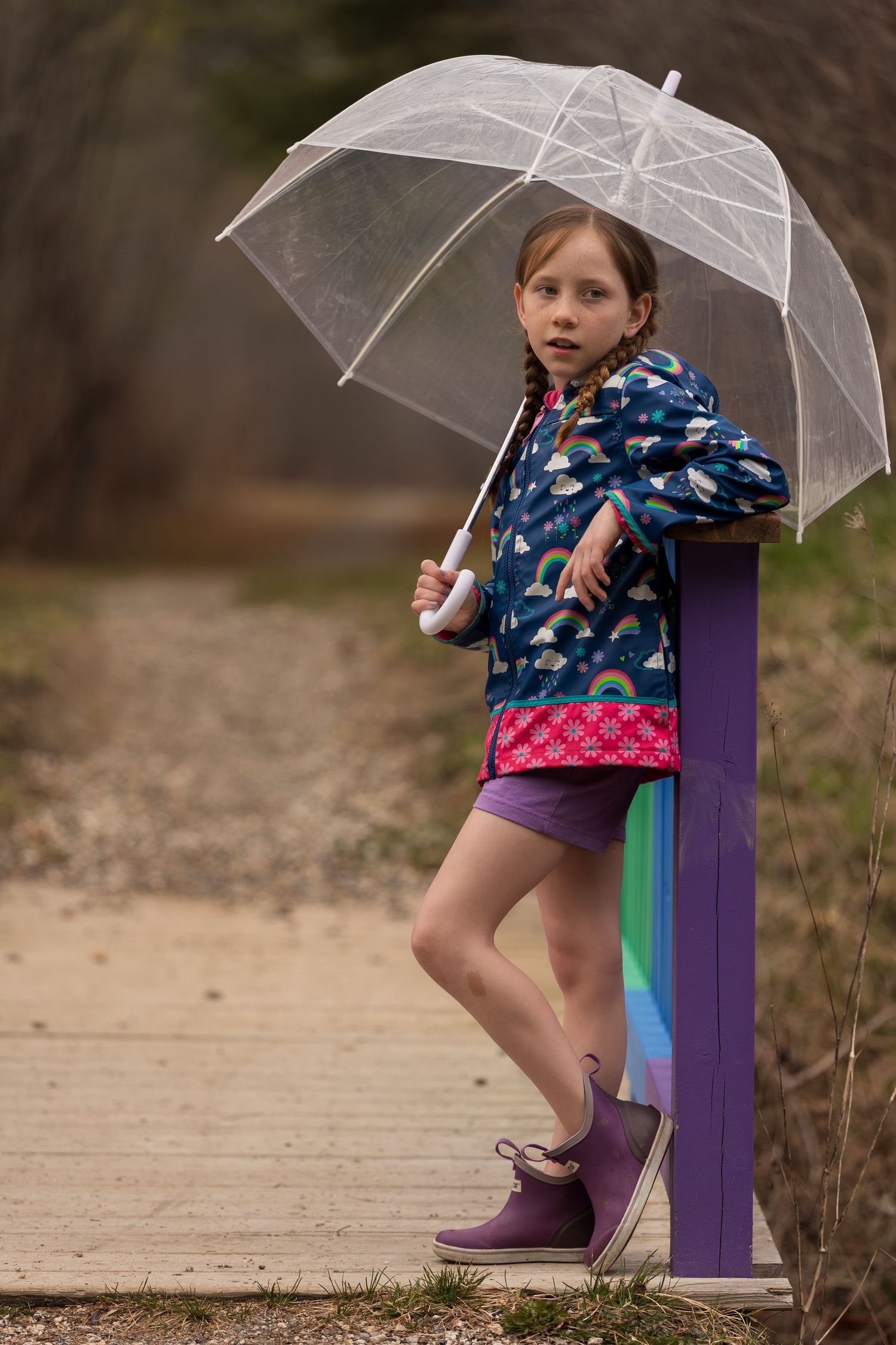 Young girl in rainbow raincoat and purple boots holds clear umbrella on wooden path.