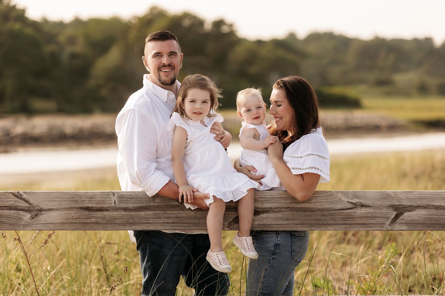 A happy family poses together on a wooden fence at sunset in a golden field with tall grass.