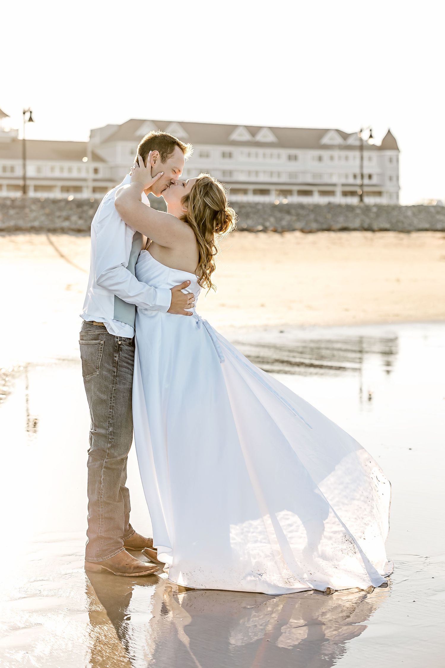 A couple shares a romantic kiss on a sandy beach at sunset while the bride's white dress flows in the ocean breeze.