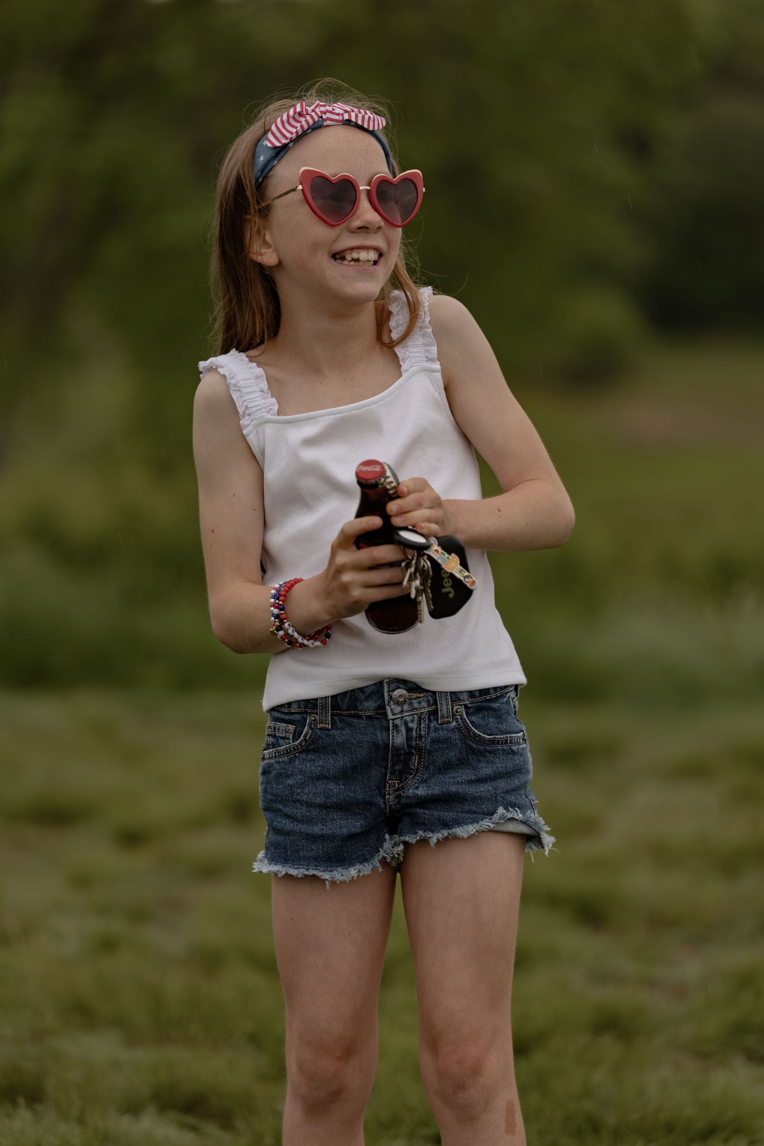 Young girl in heart sunglasses and white tank top holding bottles outdoors in a green field.