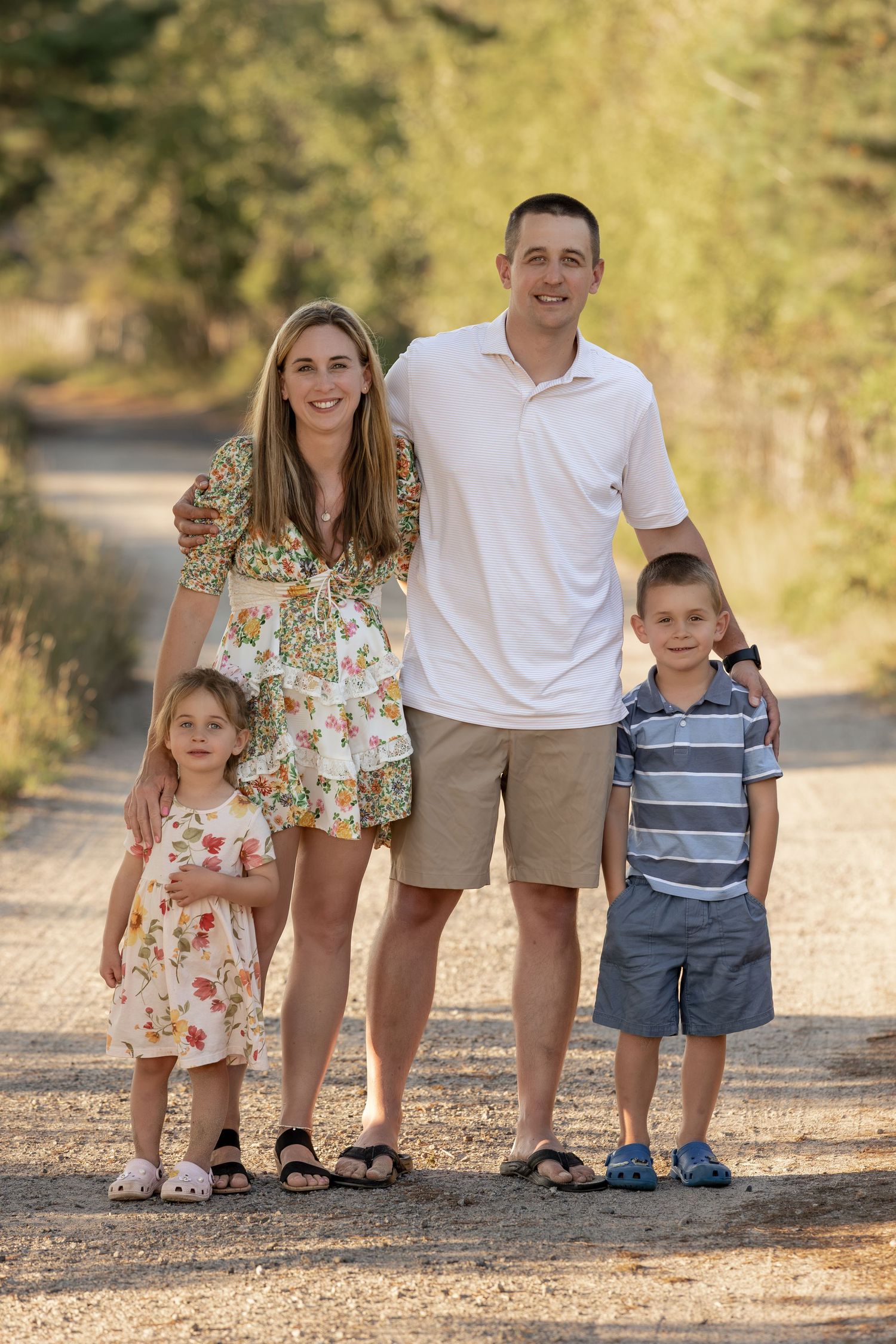 Happy family of four posing on a scenic gravel path surrounded by lush green trees on a sunny day.