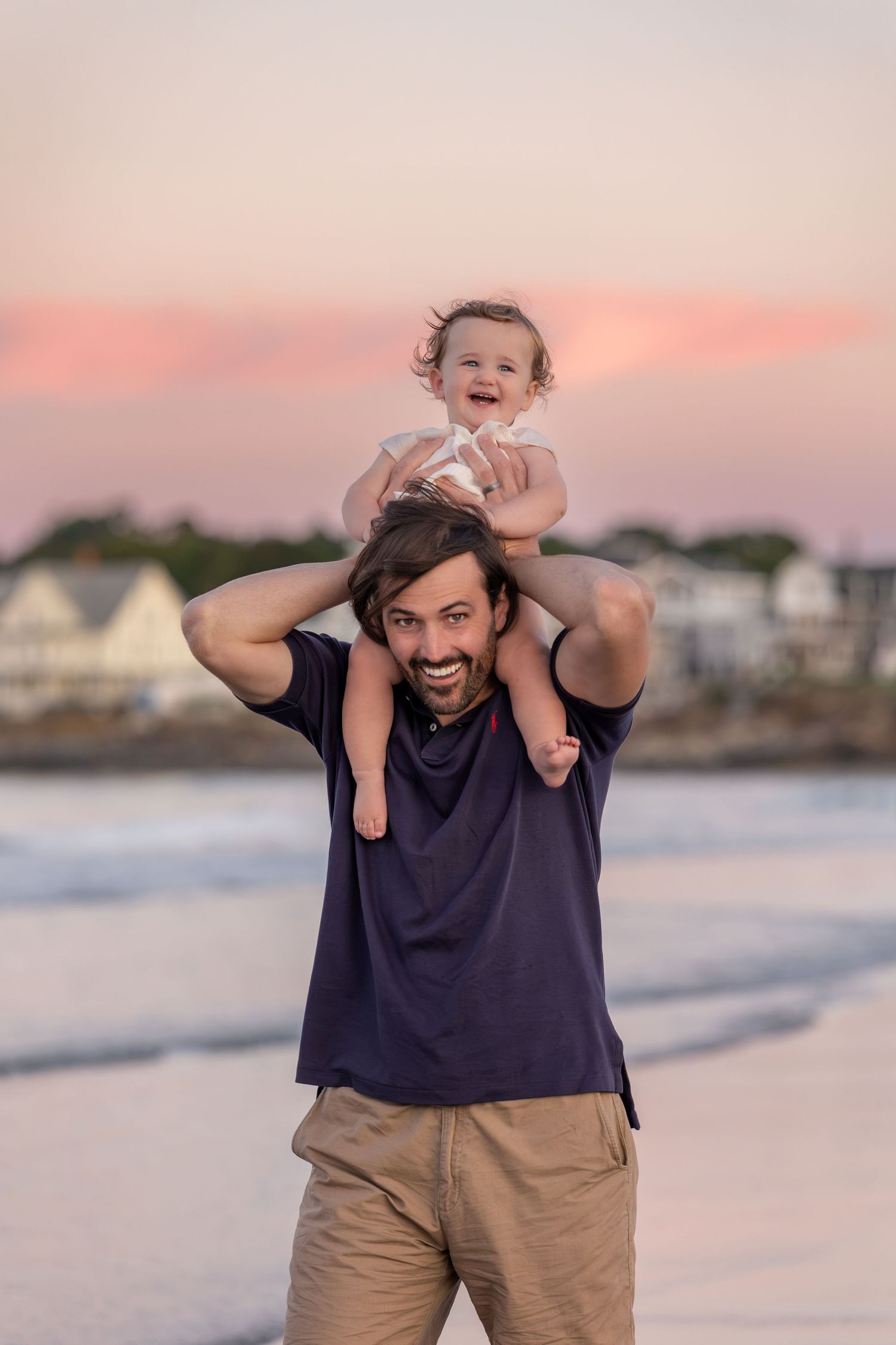 A smiling man carries a laughing baby on his shoulders at a beach during a pink sunset.
