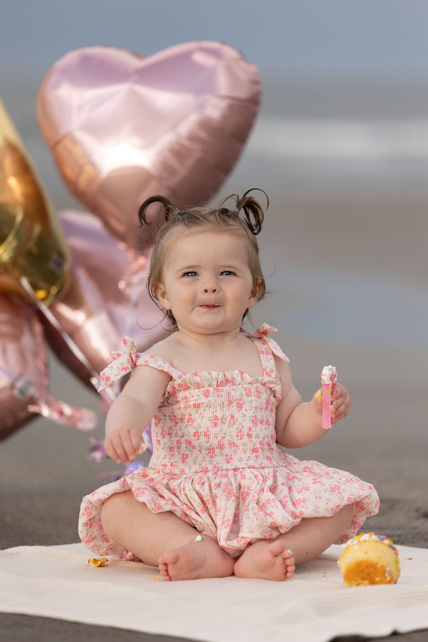 Baby girl in floral dress sits on beach blanket with heart balloons, holding cake pop at first birthday celebration.