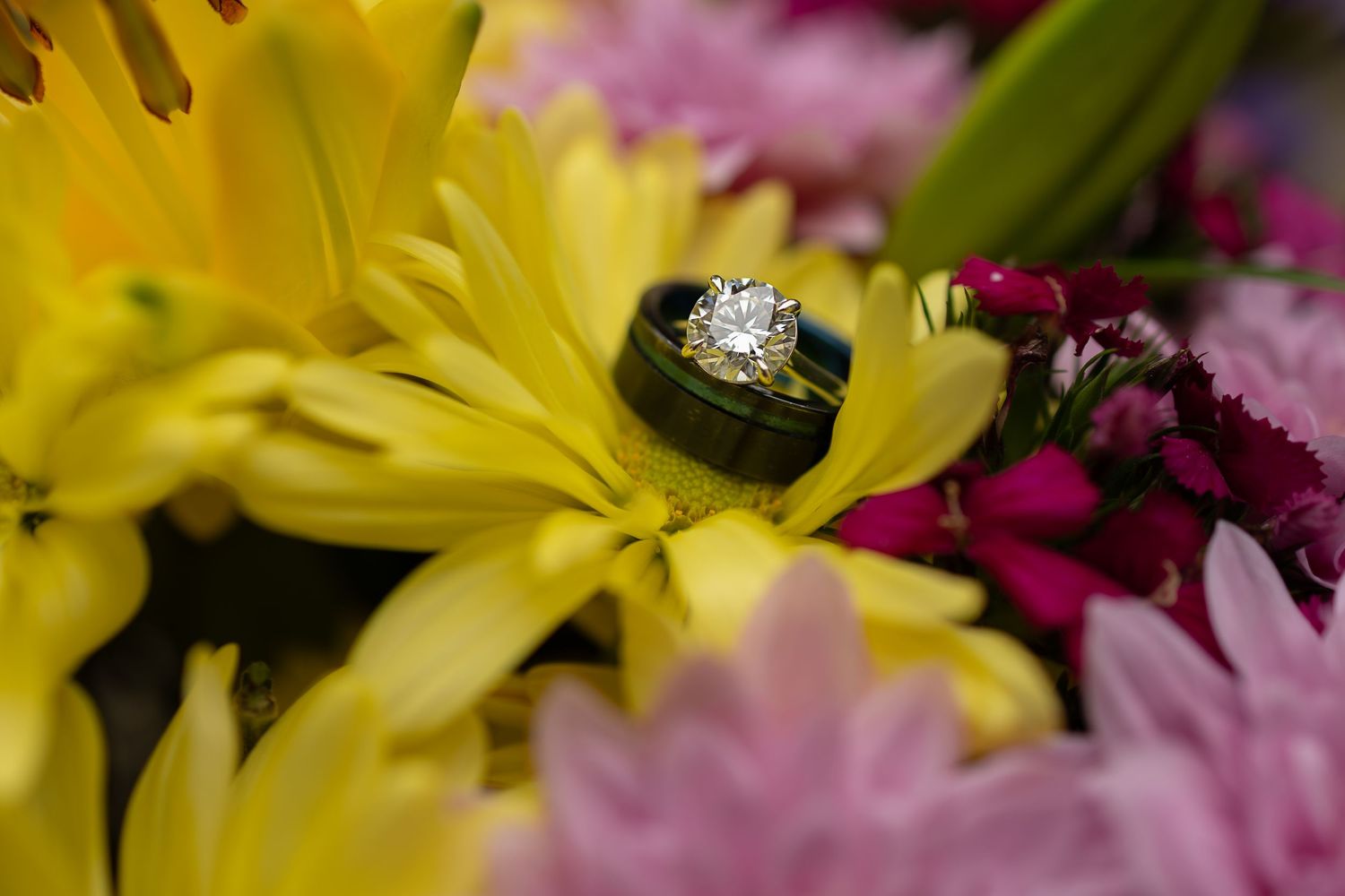 Diamond engagement ring and gold wedding band resting on vibrant yellow, pink, and purple flowers in a bridal bouquet.