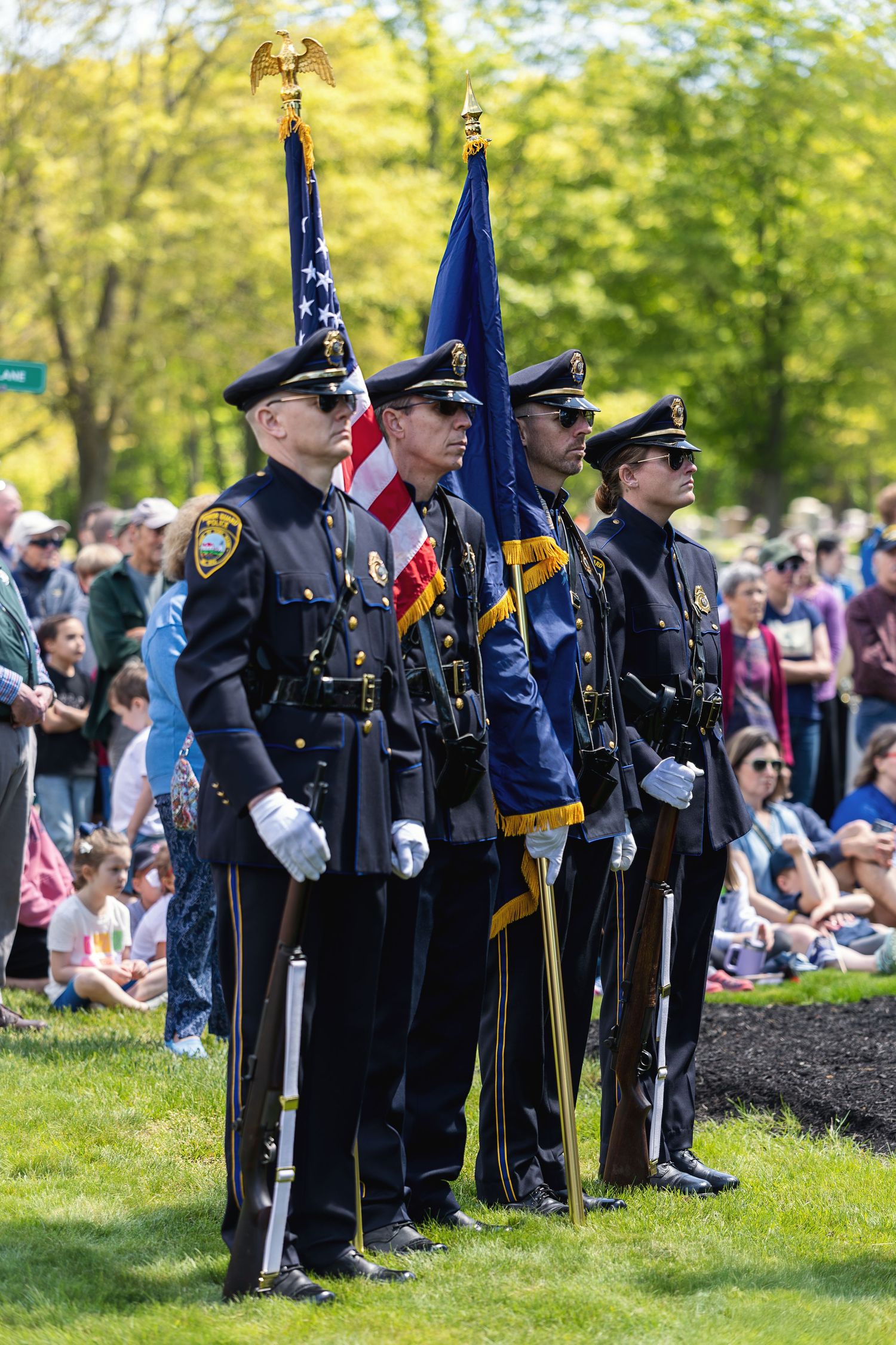Police honor guard in dress uniforms carrying American and blue flags at an outdoor ceremony with crowd.