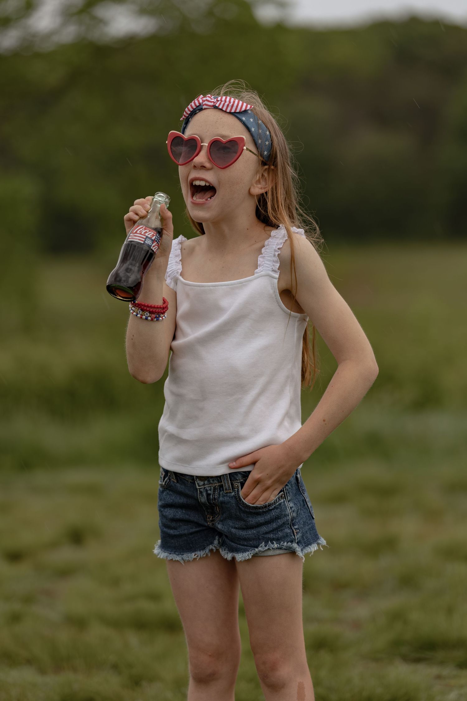 Happy girl in heart sunglasses holding a Coca-Cola bottle while laughing outdoors.
