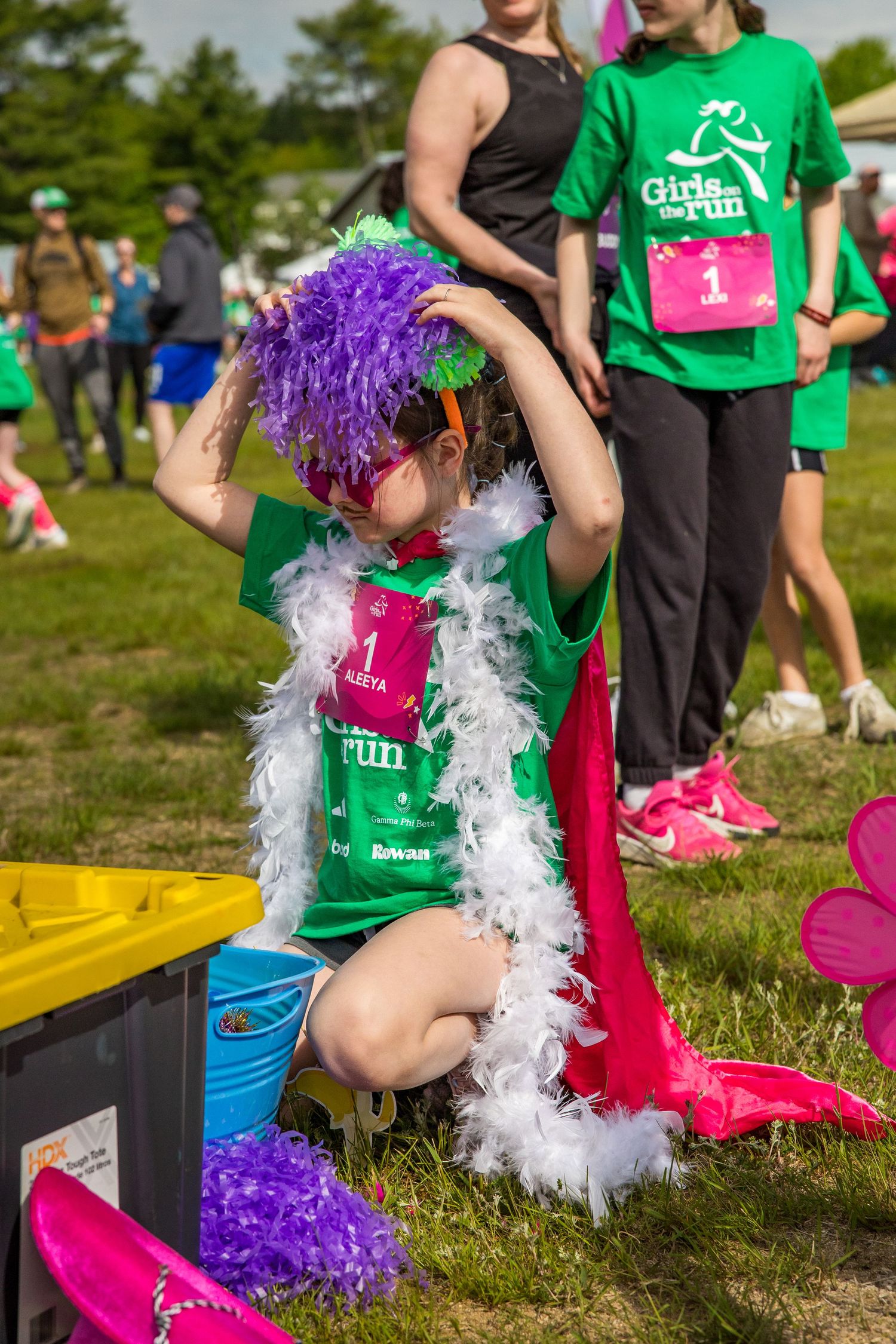 Young girl in green Girls on the Run shirt, purple wig, and feather boa adjusts costume at charity race event.