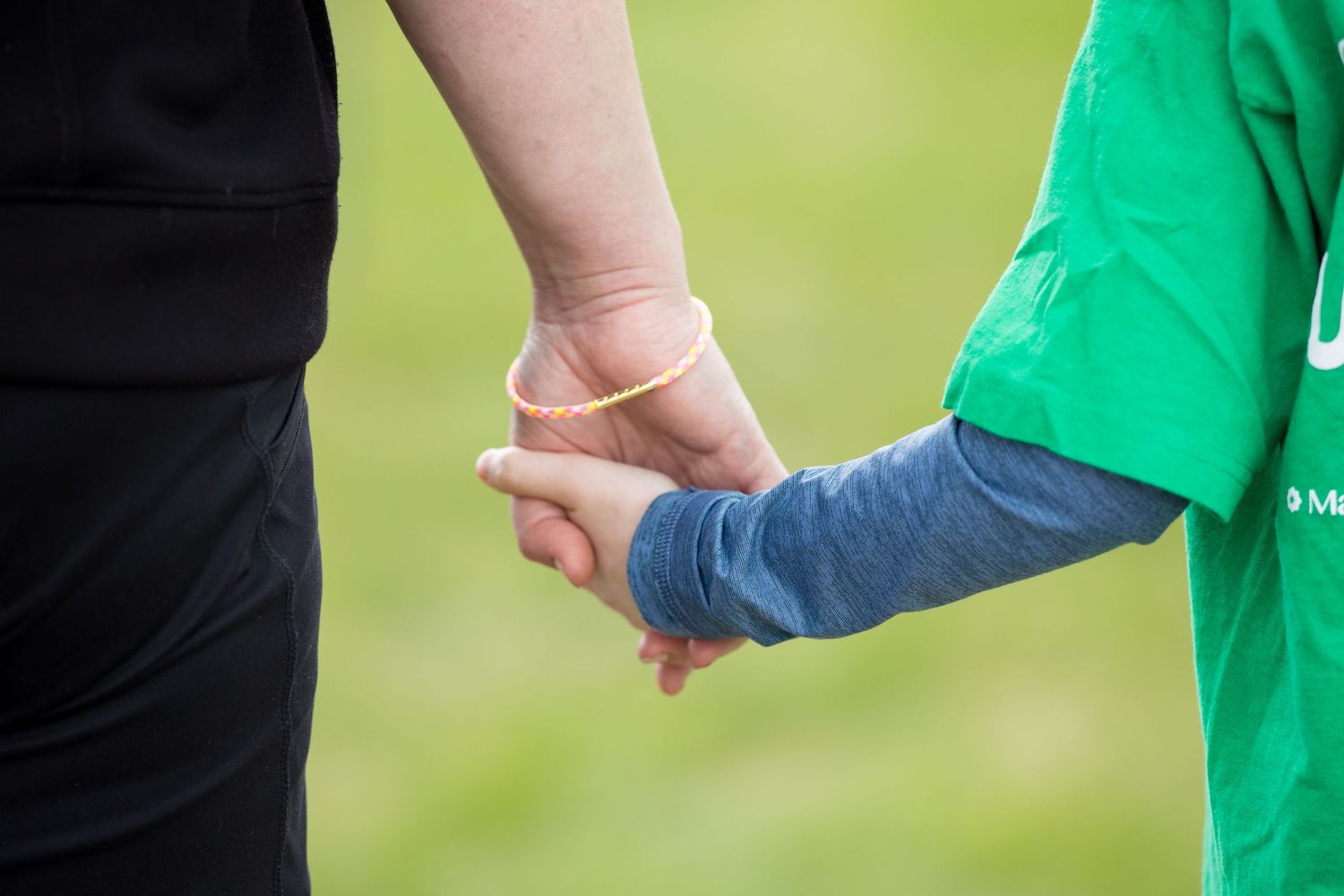 Adult and child holding hands outdoors, wearing black and green shirts, with a rubber band on the adult's wrist.